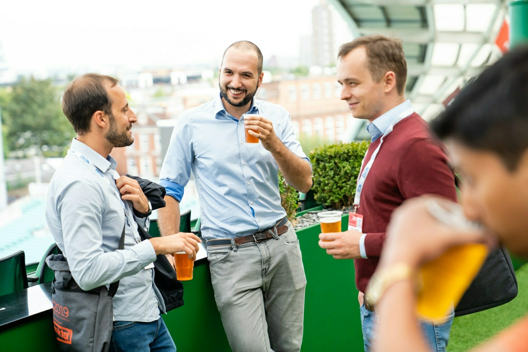 Four men are socializing on a rooftop, holding cups of beer. They are casually dressed, smiling, and appear to be enjoying a lively conversation. The background includes greenery and a city skyline. James Gifford-Mead Photography - Event Photographer London