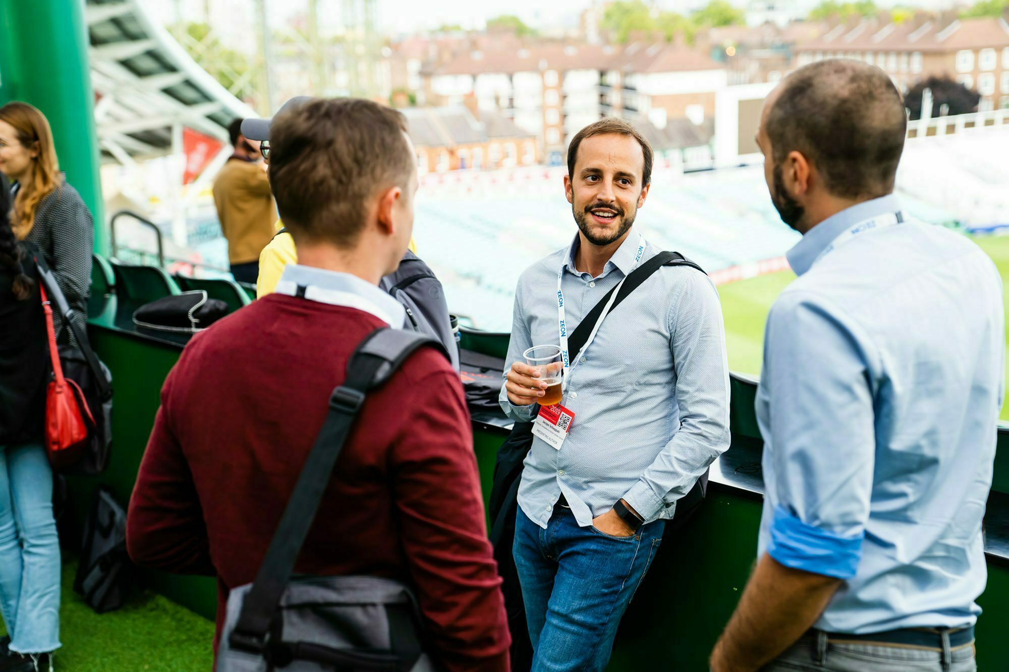 A group of men stand outside at a stadium, casually talking and holding drinks. They are wearing casual clothing with ID lanyards. One man holds a glass and smiles. A green field and stadium seating are visible in the background. James Gifford-Mead Photography - Event Photographer London