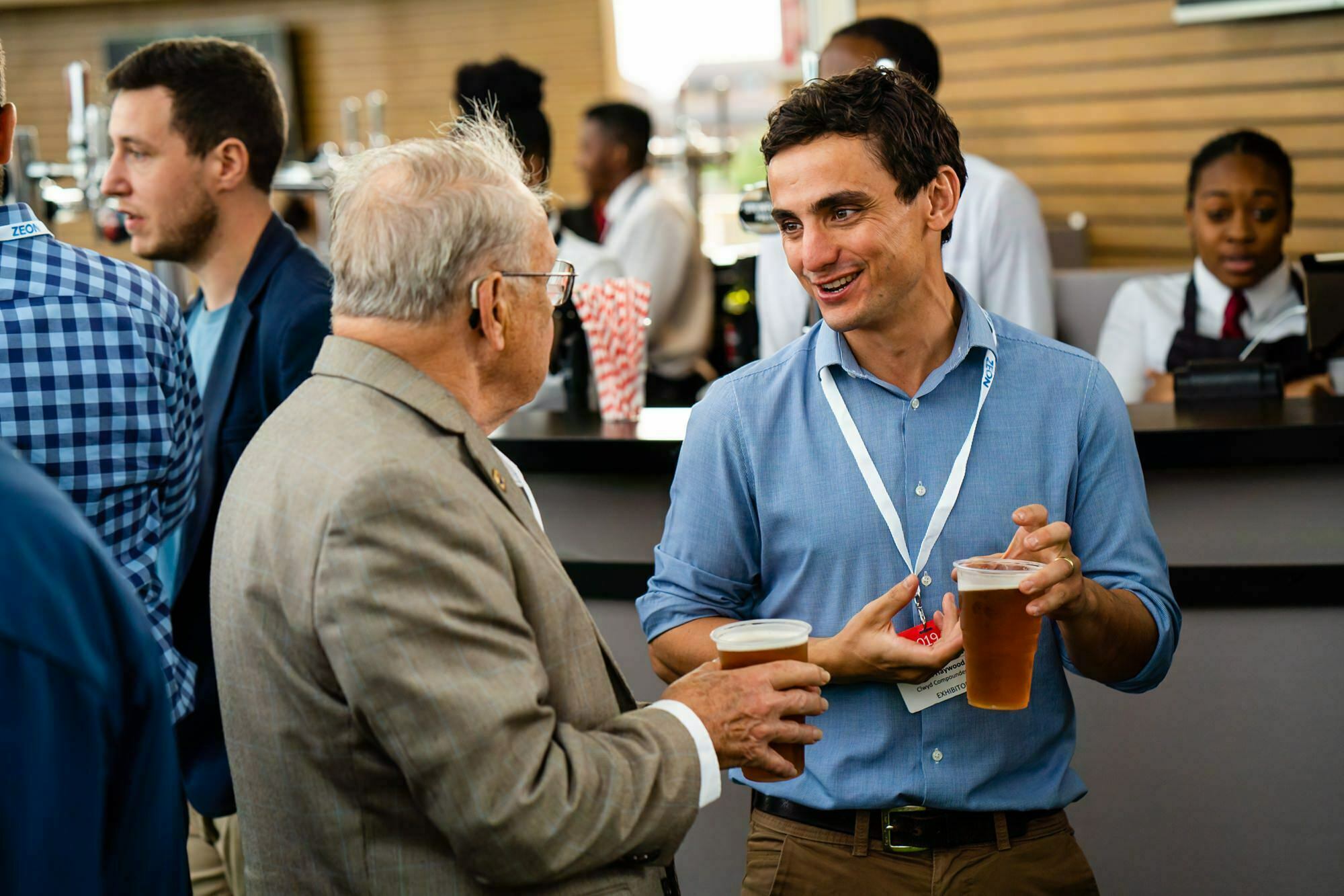 Two men are standing and talking while holding drinks at an event. The older man is wearing a light suit, and the younger man is in a blue shirt with a lanyard. Other attendees and staff are visible in the background. James Gifford-Mead Photography - Event Photographer London