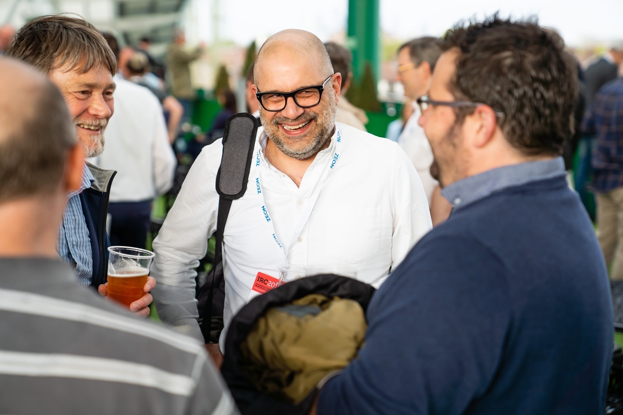 A group of people are gathered outdoors, smiling and talking. A man in the center wearing glasses holds a backpack and has a conference badge around his neck. Others around him hold drinks. The setting appears informal and social. James Gifford-Mead Photography - Event Photographer London