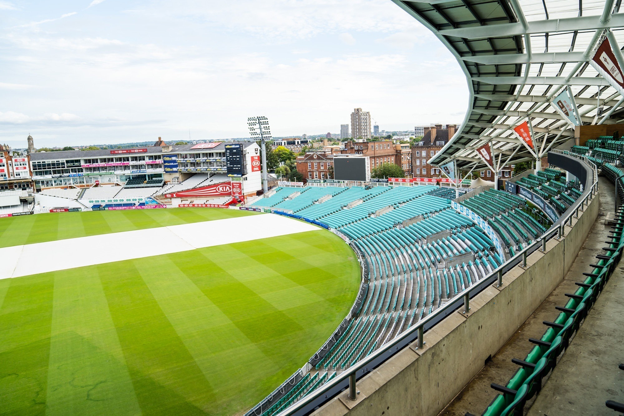 A panoramic view of an empty sports stadium with green seating and a well-maintained grass field. The sky is partly cloudy, and the cityscape is visible in the background. The stadium appears ready for a sporting event. James Gifford-Mead Photography - Event Photographer London