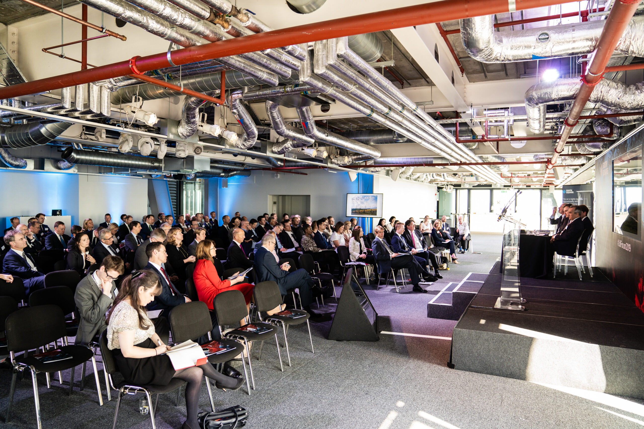 A conference room with a large audience seated and facing a panel of speakers on a stage. The room has visible industrial-style piping and large windows allowing natural light. Attendees are dressed in formal business attire. James Gifford-Mead Photography - Event Photographer London