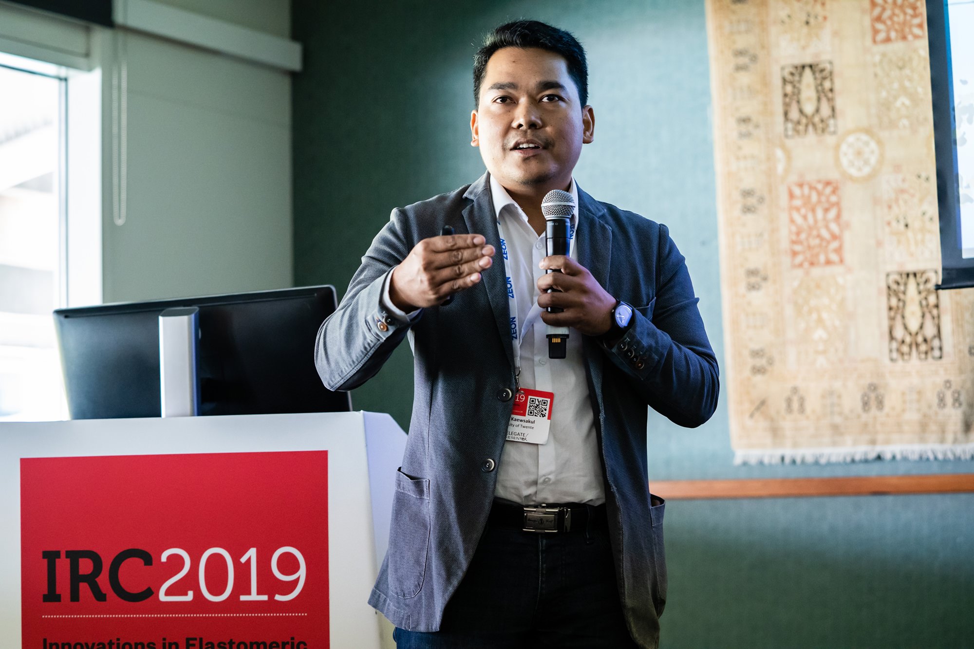 A person holding a microphone and speaking at a podium with "IRC 2019 Innovations in Elastomeric" displayed on the front. The individual is wearing a grey blazer, standing in a room with green walls and a patterned wall hanging. James Gifford-Mead Photography - Event Photographer London
