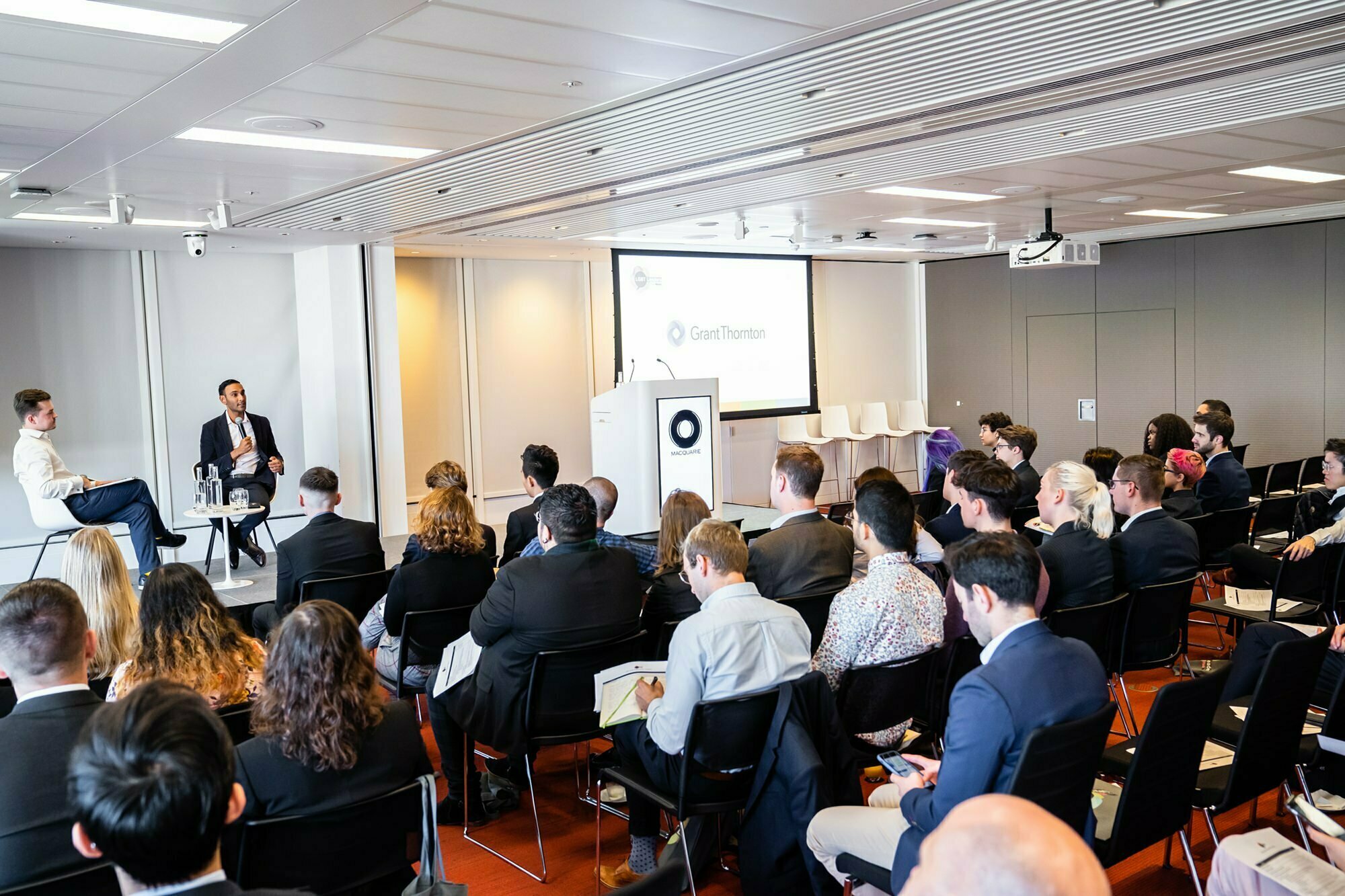 A conference room filled with attendees seated in rows facing a stage. Two presenters sit and converse on a panel discussion. A screen behind them displays a presentation slide with the Grant Thornton logo. James Gifford-Mead Photography - Event Photographer London