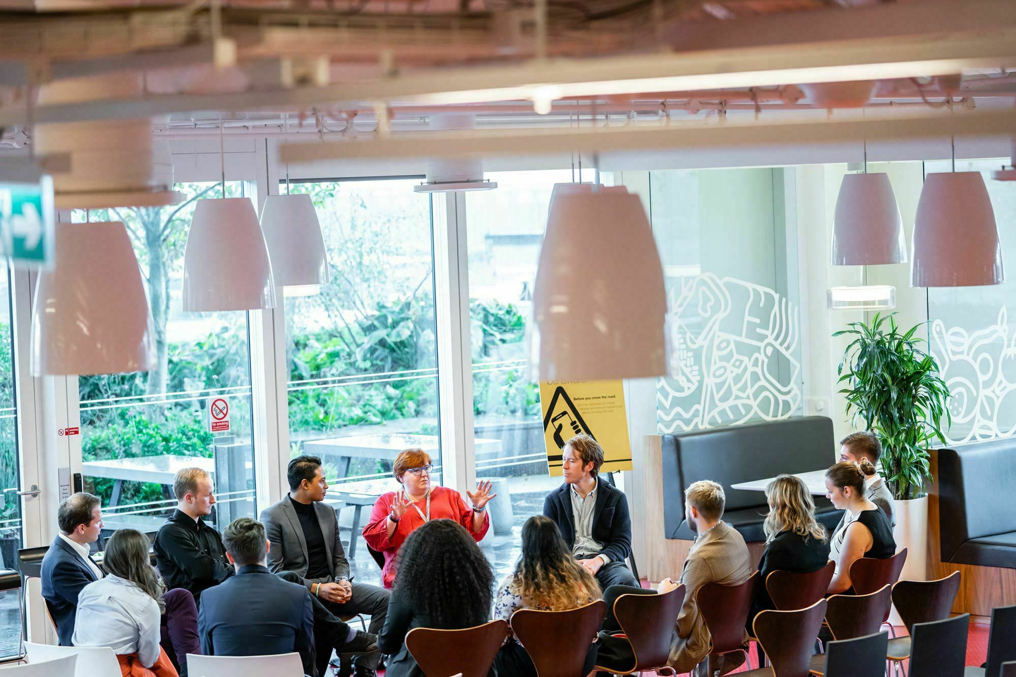 A group of people sit in a circle having a discussion in a bright room with large windows. The setting features modern decor with hanging lamps and a plant in the corner. One person is speaking while others listen attentively. James Gifford-Mead Photography - Event Photographer London