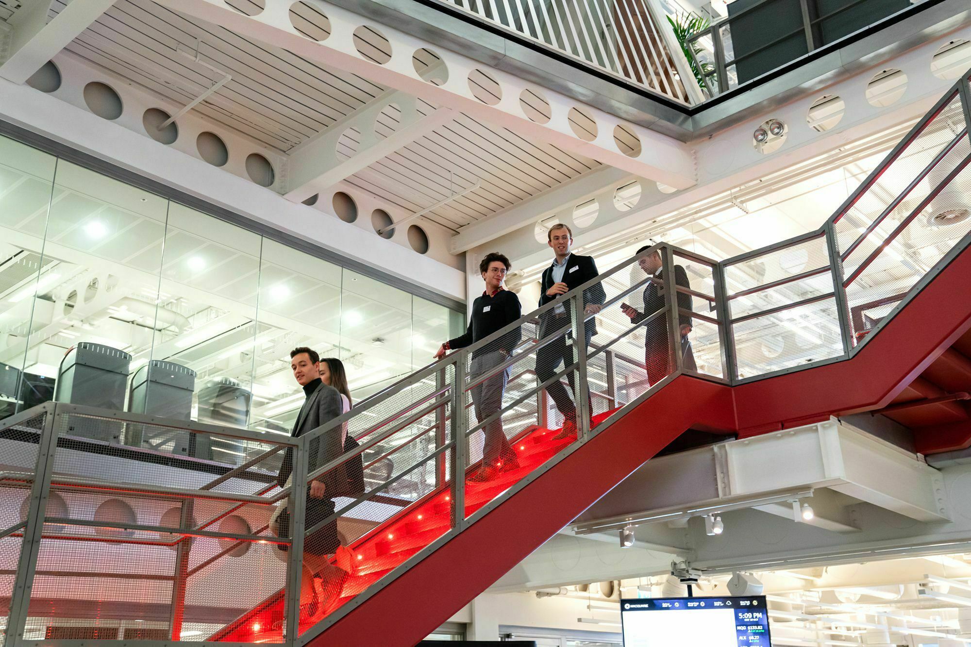 A group of business professionals in suits descend a red staircase inside a modern office with glass walls and exposed beams. Fluorescent lighting and a digital screen are visible in the background. James Gifford-Mead Photography - Event Photographer London