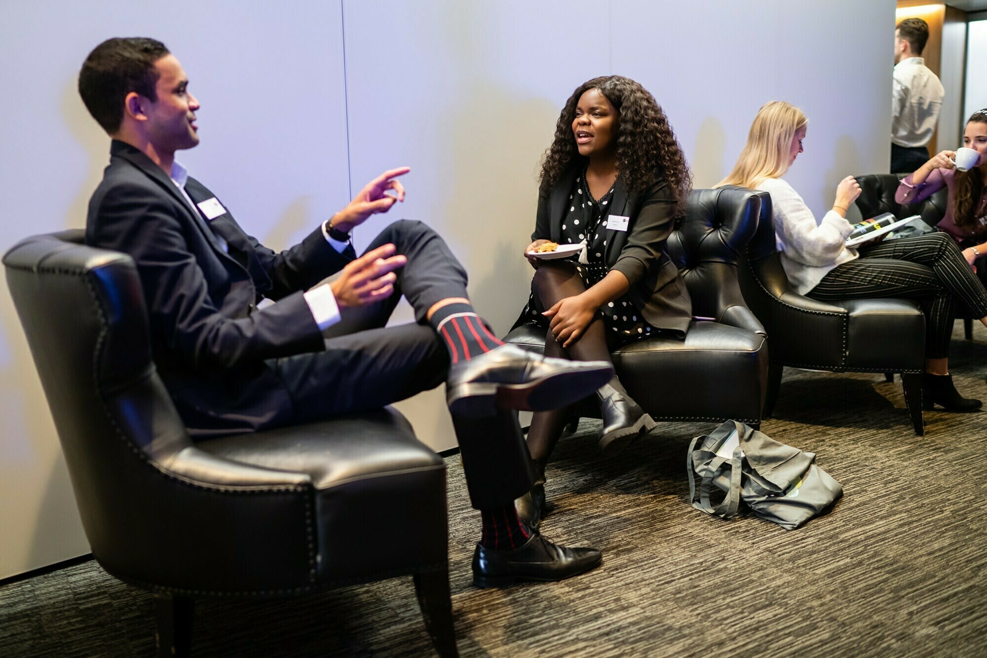 Two people in business attire sit and converse on leather chairs in a modern meeting area. A woman holds a plate, engaged in the discussion. In the background, others are seated, some eating and chatting. A bag rests on the floor nearby. James Gifford-Mead Photography - Event Photographer London