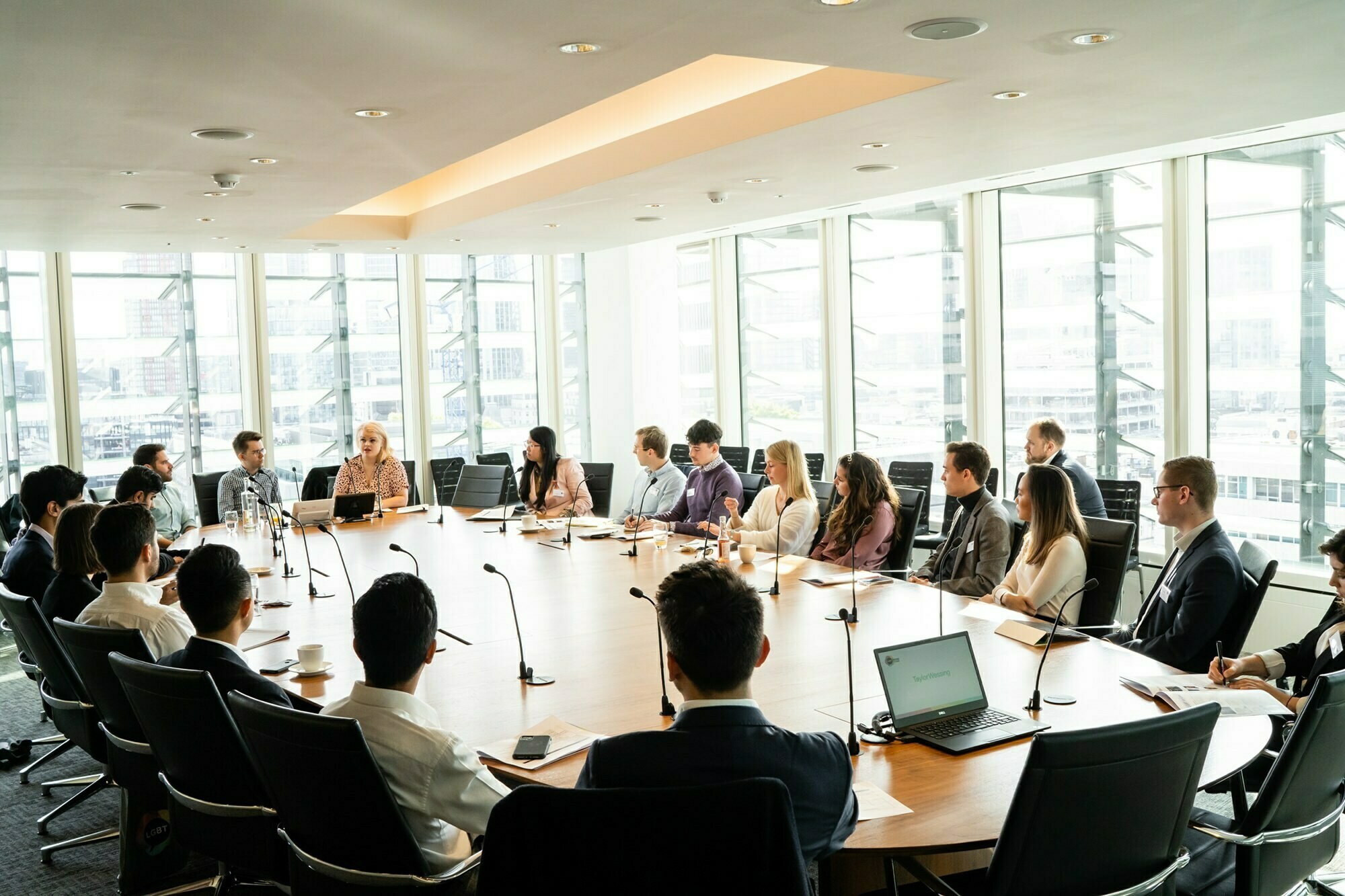 A large conference room with a long oval table surrounded by professionals engaged in a meeting. Laptops and microphones are on the table. The room features floor-to-ceiling windows, with bright natural light filling the space. James Gifford-Mead Photography - Event Photographer London