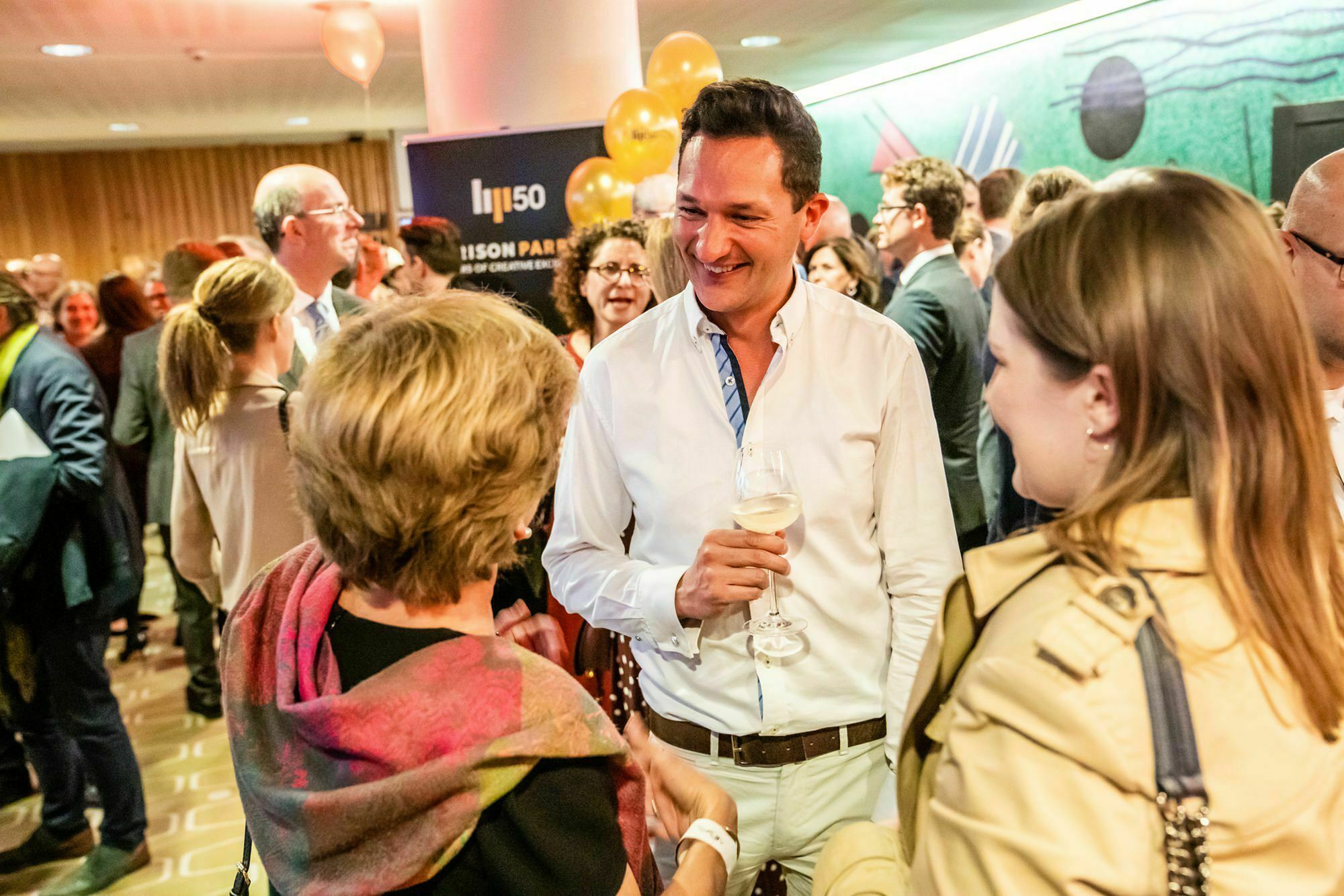 A man in a white shirt, holding a drink, smiles while talking to two women at a crowded event. People around are engaged in conversations, with balloons and a green textured wall in the background. James Gifford-Mead Photography - Event Photographer London