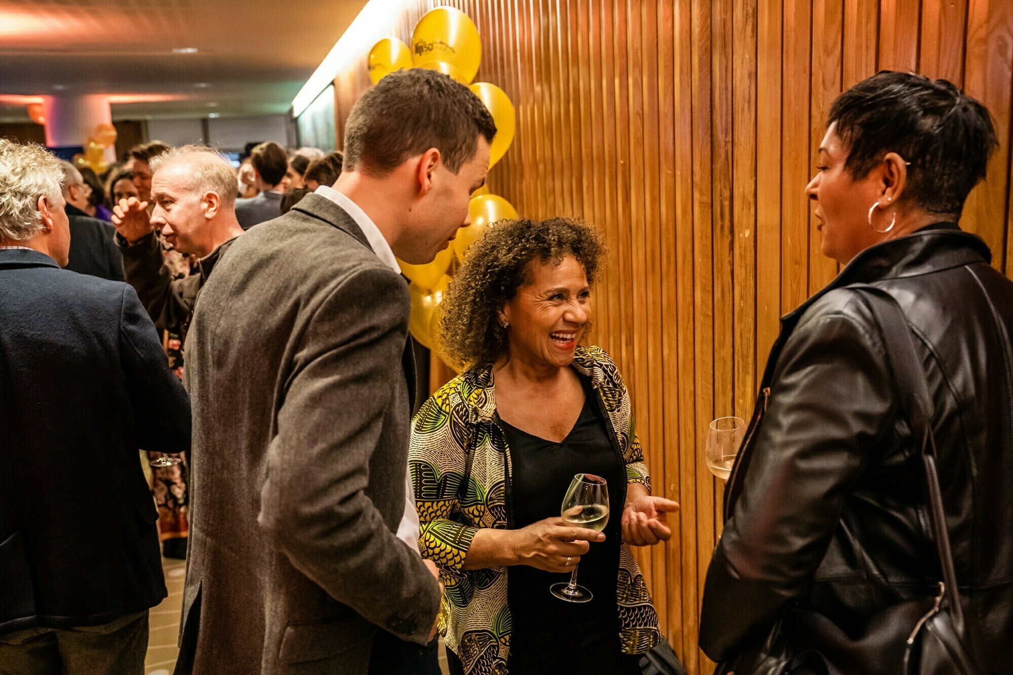 A lively social gathering with people chatting in a warmly lit room. A woman holding a wine glass is smiling while conversing with a man and another woman. Yellow balloons are in the background near a wooden wall. James Gifford-Mead Photography - Event Photographer London