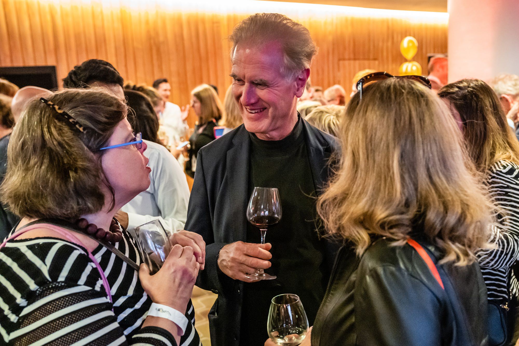 A man and two women are in conversation at a social gathering. Each holds a glass of wine. The room is filled with people and warm lighting, creating a lively and welcoming atmosphere. James Gifford-Mead Photography - Event Photographer London