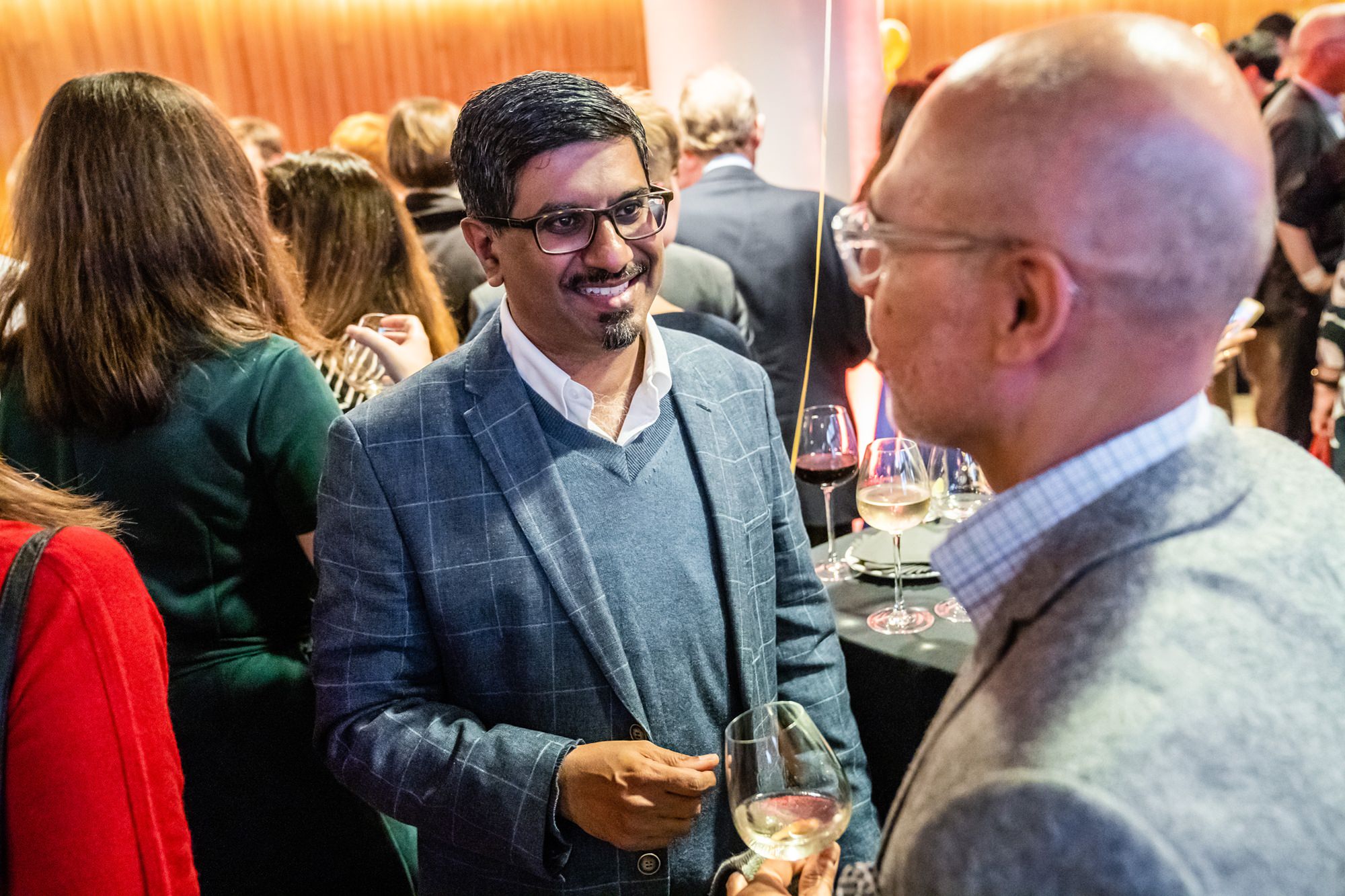 Two men in business attire are engaged in conversation at a lively social event. They are holding glasses of wine. The background is filled with people mingling, and the setting has warm lighting. James Gifford-Mead Photography - Event Photographer London