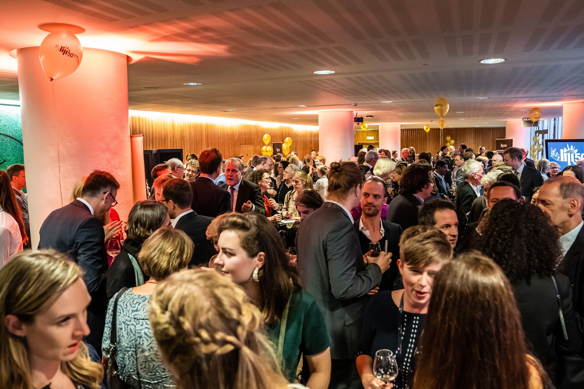 A large group of people in formal attire gather and socialize at an indoor event. Balloons and soft lighting create a festive atmosphere. James Gifford-Mead Photography - Event Photographer London