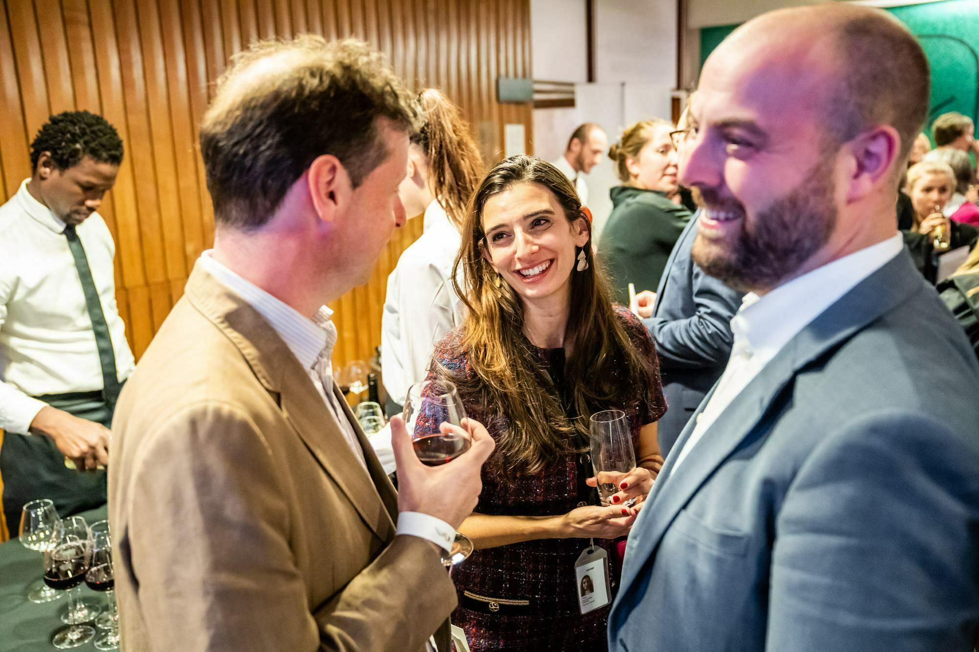 Three people are engaged in conversation at a social event with wine glasses in hand. The background shows more attendees and a waiter. The setting appears lively and convivial, with warm lighting and wooden decor. James Gifford-Mead Photography - Event Photographer London