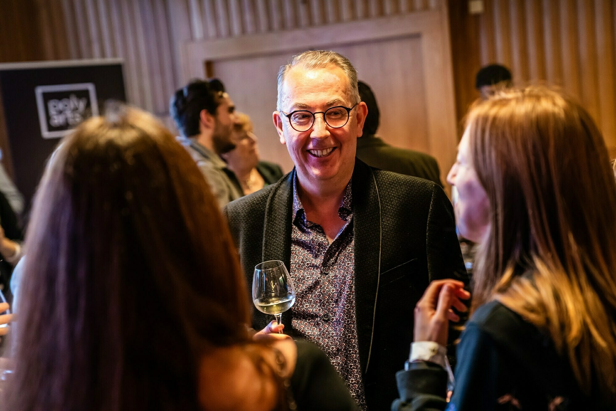 A man in a dark blazer and glasses smiles while holding a glass of white wine, engaging with two women in a lively indoor setting. The scene is warmly lit, with other guests mingling in the background. James Gifford-Mead Photography - Event Photographer London