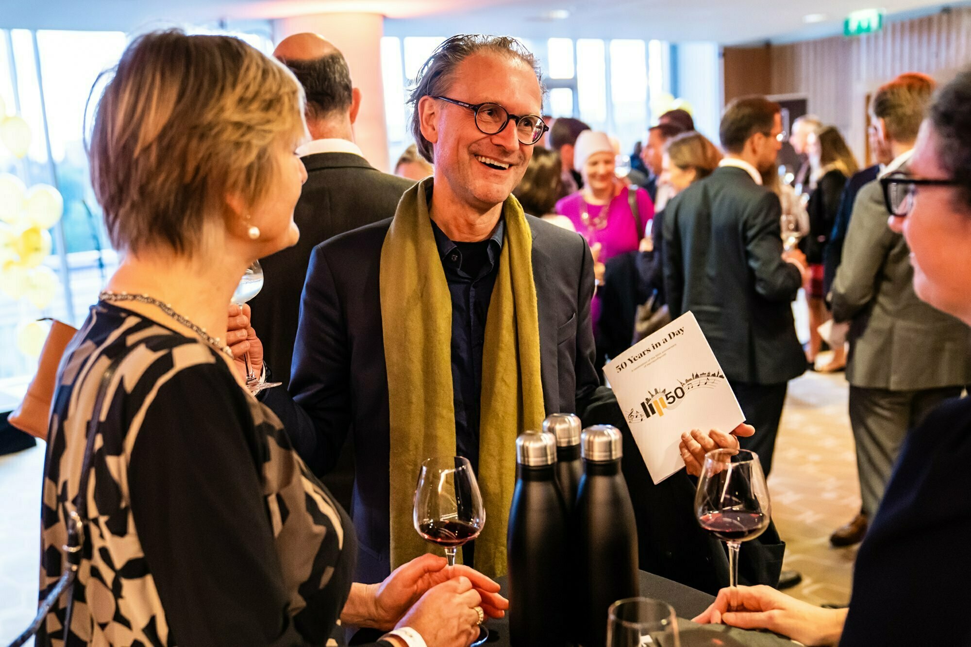 A group of people in formal attire are socializing at an event. One man, wearing glasses and a green scarf, is smiling and holding a program. Wine glasses and bottles are on the table. They stand in a well-lit room with a large window. James Gifford-Mead Photography - Event Photographer London