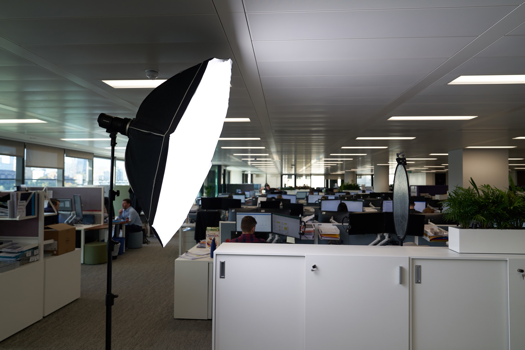 A large softbox light is set up in an open-plan office with numerous desks and monitors. People are working in the background. The office is well-lit, with a mix of artificial and natural light coming through the windows. James Gifford-Mead Photography - Event Photographer London