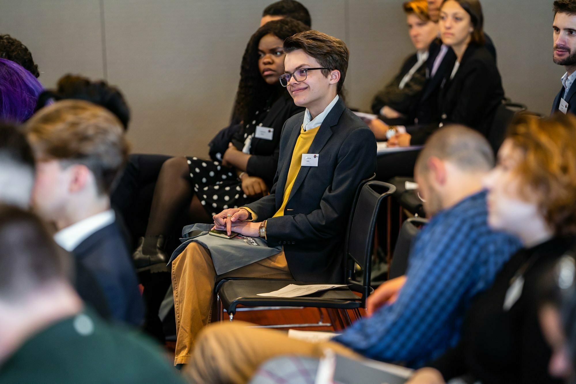 A group of people seated in a conference room, attentively listening. A person in a suit and glasses smiles, holding a notepad. Others around them are focused forward. Name tags are visible on some attendees. James Gifford-Mead Photography - Event Photographer London