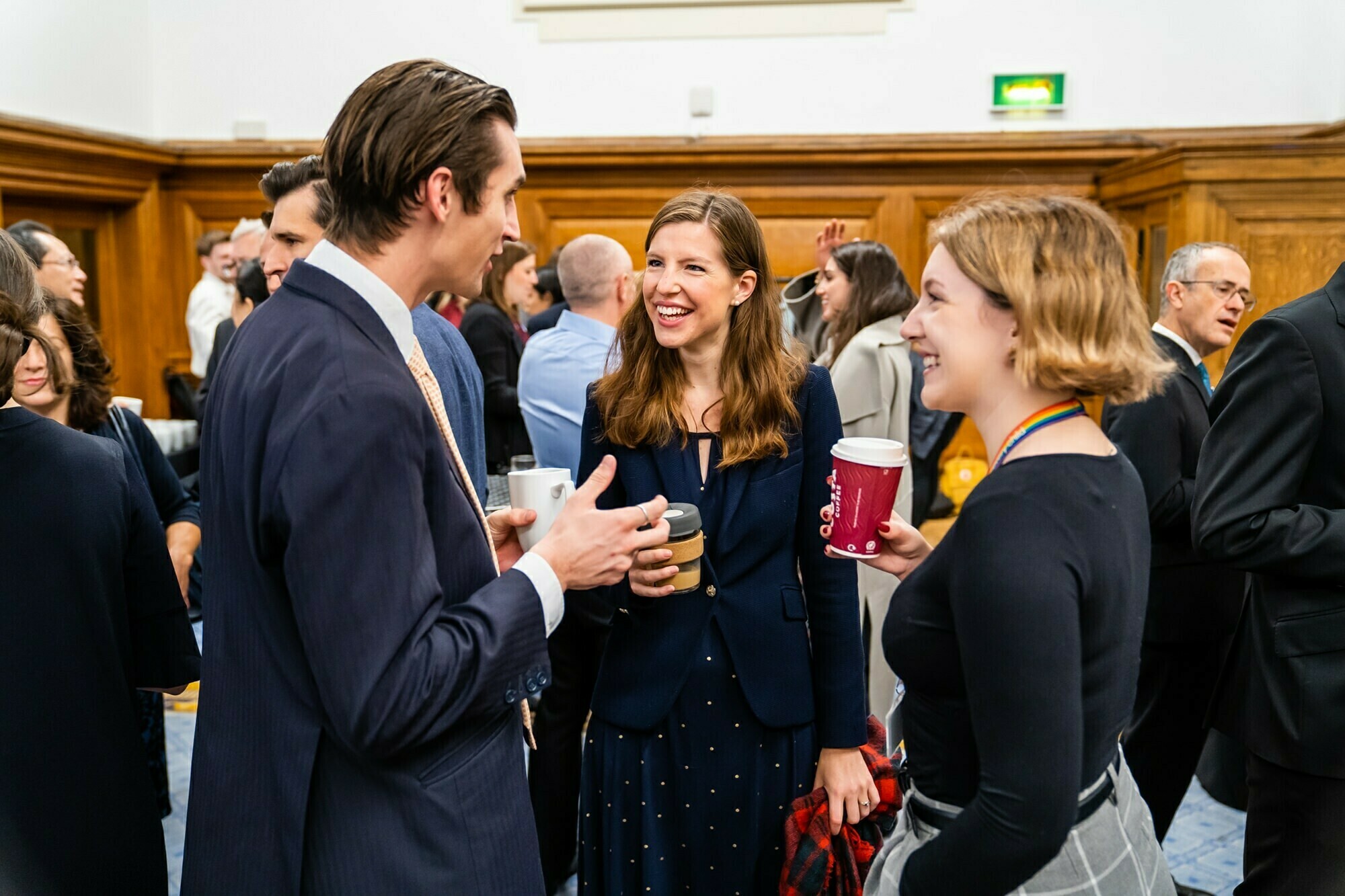 A group of people in business attire are engaged in conversation, holding coffee cups. A woman in the center is smiling and listening to a man speaking. They are in a room with wood-paneled walls, surrounded by other mingling individuals. James Gifford-Mead Photography - Event Photographer London