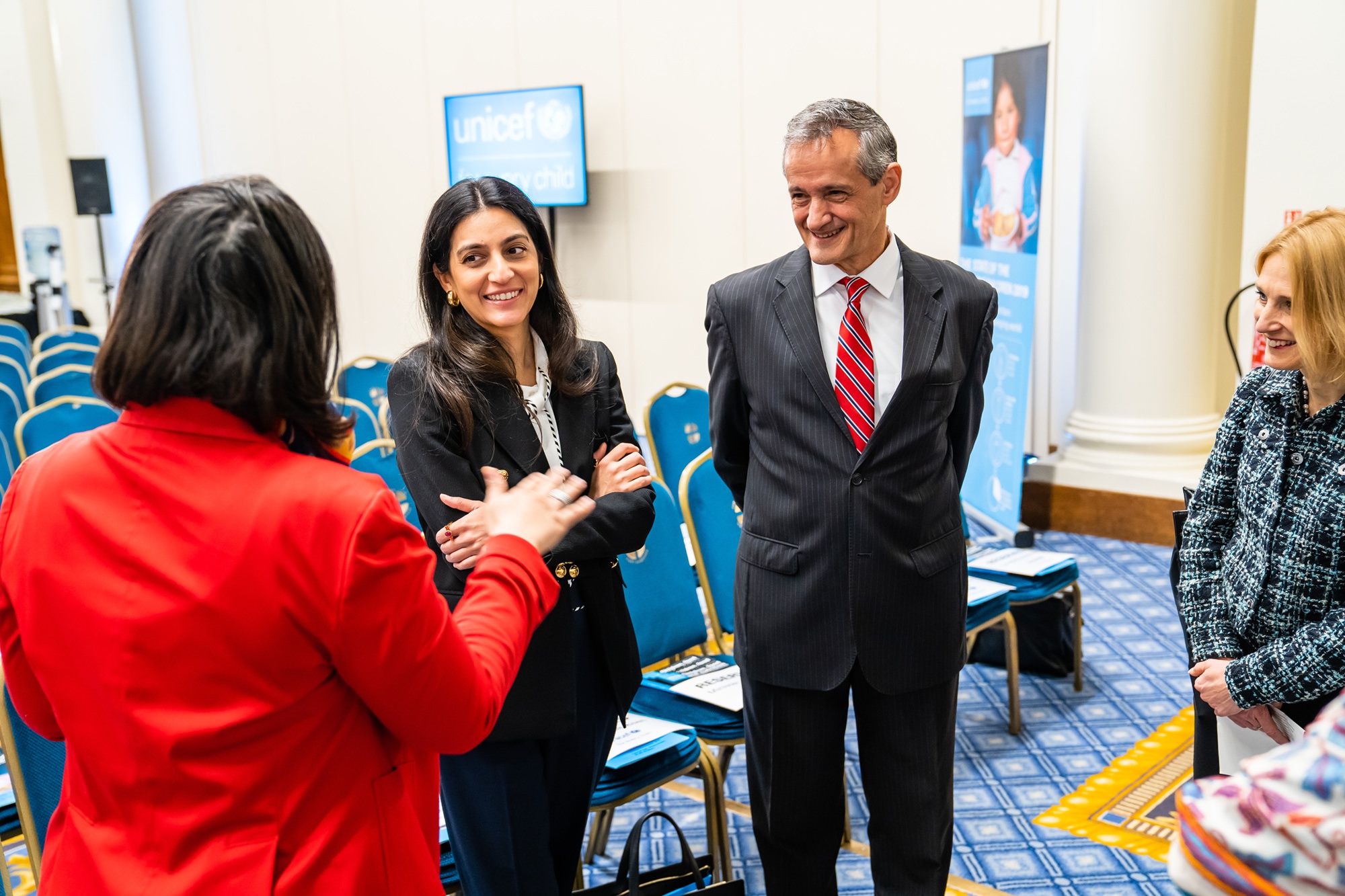 Four people are engaged in conversation in a conference room with UNICEF posters in the background. Three are standing together, attentively listening to the fourth person, who is gesturing while speaking. Blue chairs and patterned carpet are visible. James Gifford-Mead Photography - Event Photographer London