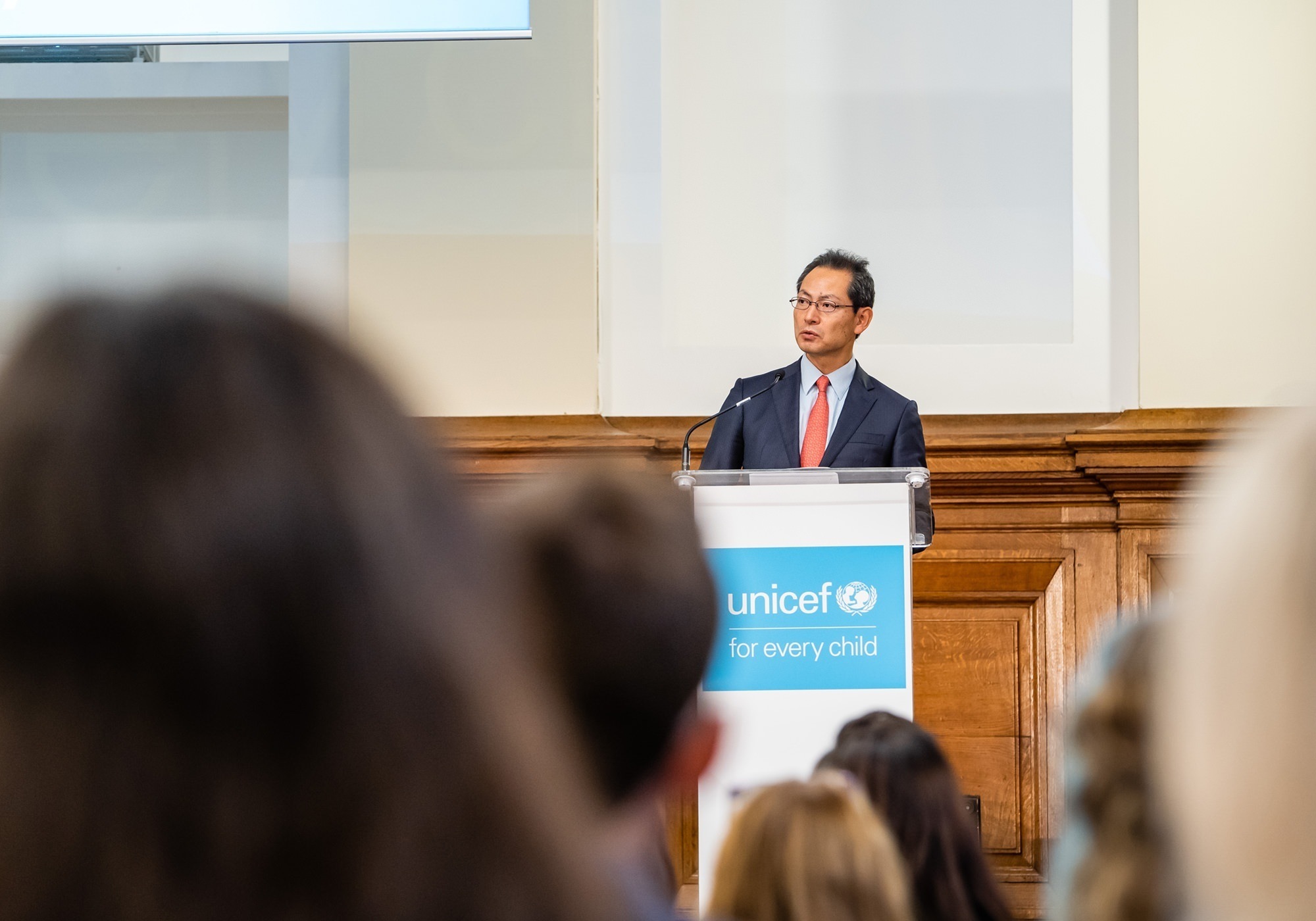 A man in a suit and red tie speaks at a podium with a UNICEF logo. The audience is partially visible in the foreground. The room has wooden paneling and a screen in the background. James Gifford-Mead Photography - Event Photographer London