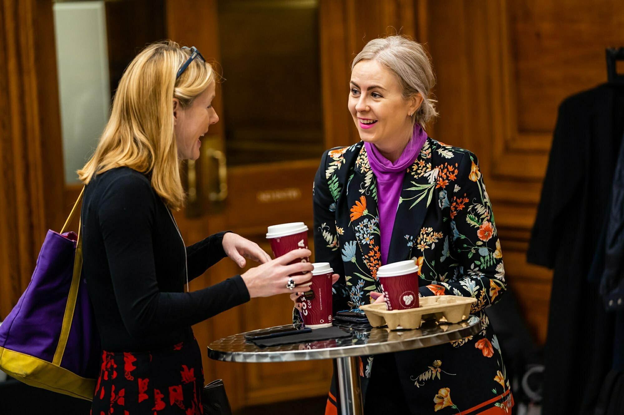 Two women converse at a small round table, each holding a coffee cup. One wears a floral blazer and a purple turtleneck, while the other sports a black top and red skirt. They appear engaged and smiling, with a coat rack and wooden door in the background. James Gifford-Mead Photography - Event Photographer London