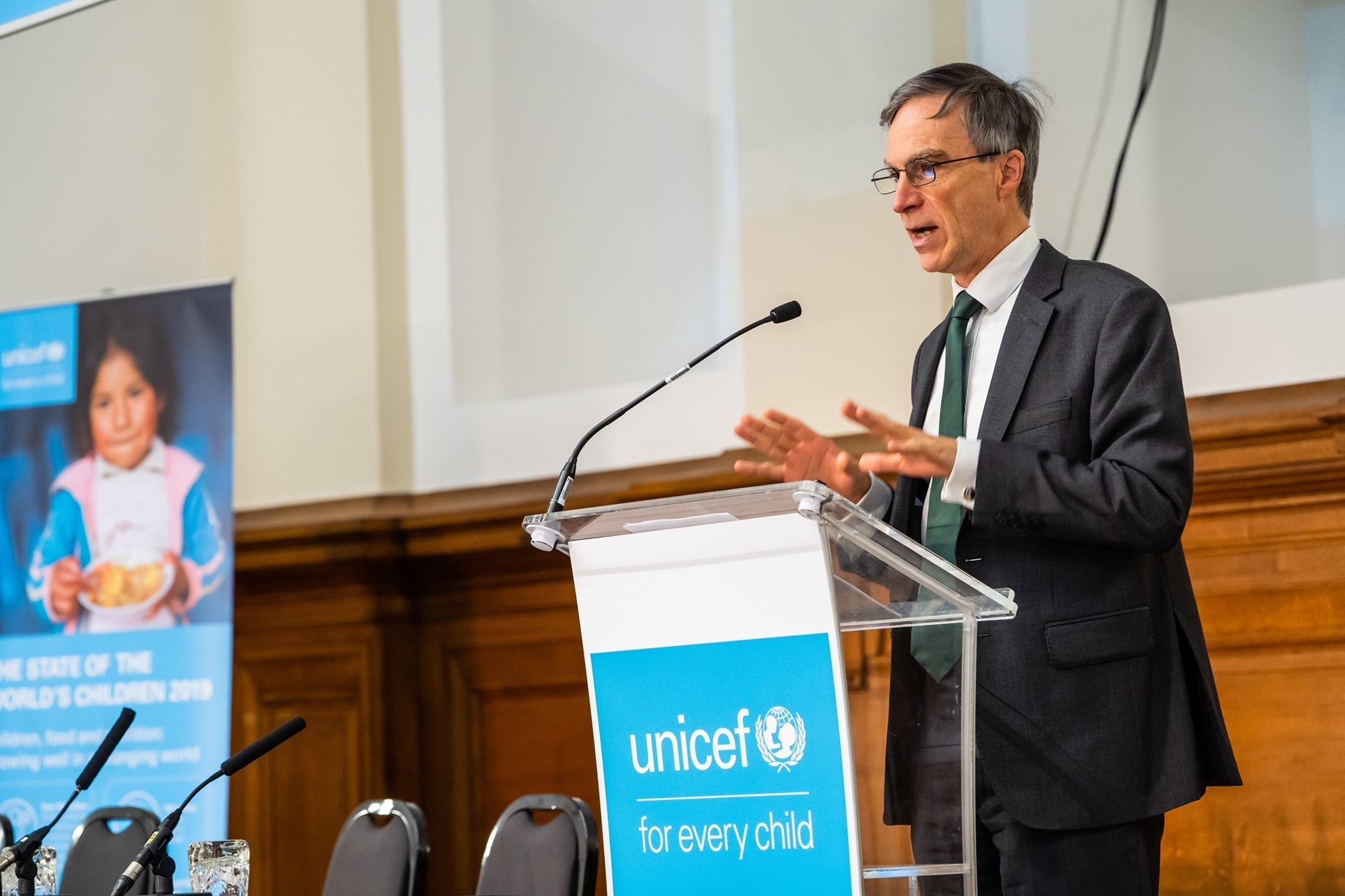 A man in a suit speaks at a podium displaying the UNICEF logo. Behind him, a poster features a child and mentions "The State of the World's Children 2019." The setting appears to be a formal conference room. James Gifford-Mead Photography - Event Photographer London
