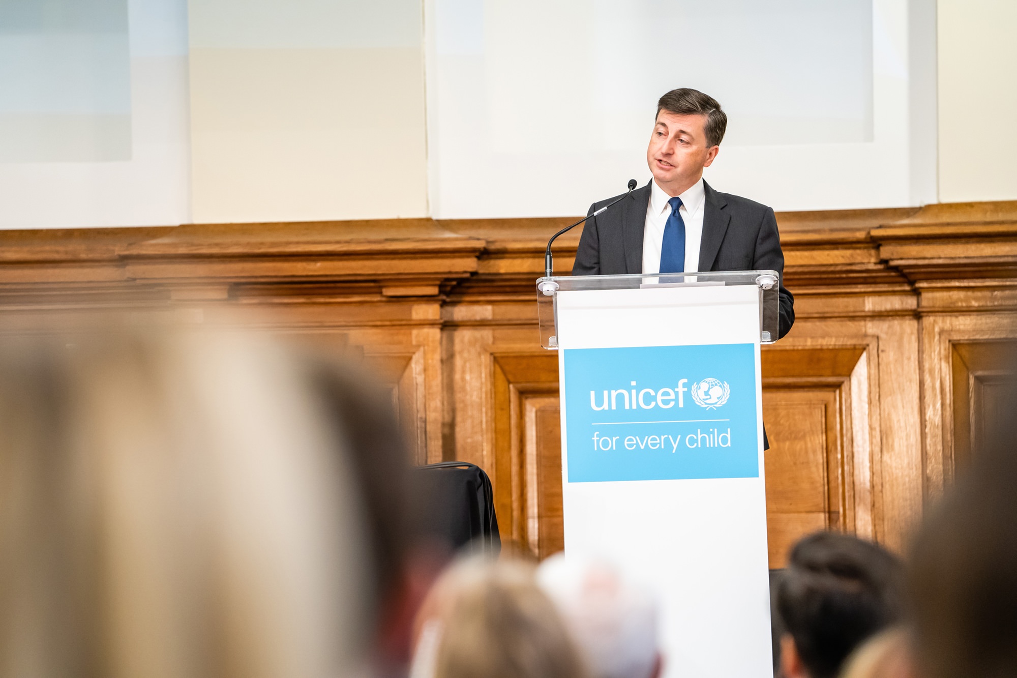 A man in a suit stands behind a podium with a UNICEF logo and the text "for every child." He is speaking in a room with wooden paneling. Audience members are visible in the foreground. James Gifford-Mead Photography - Event Photographer London