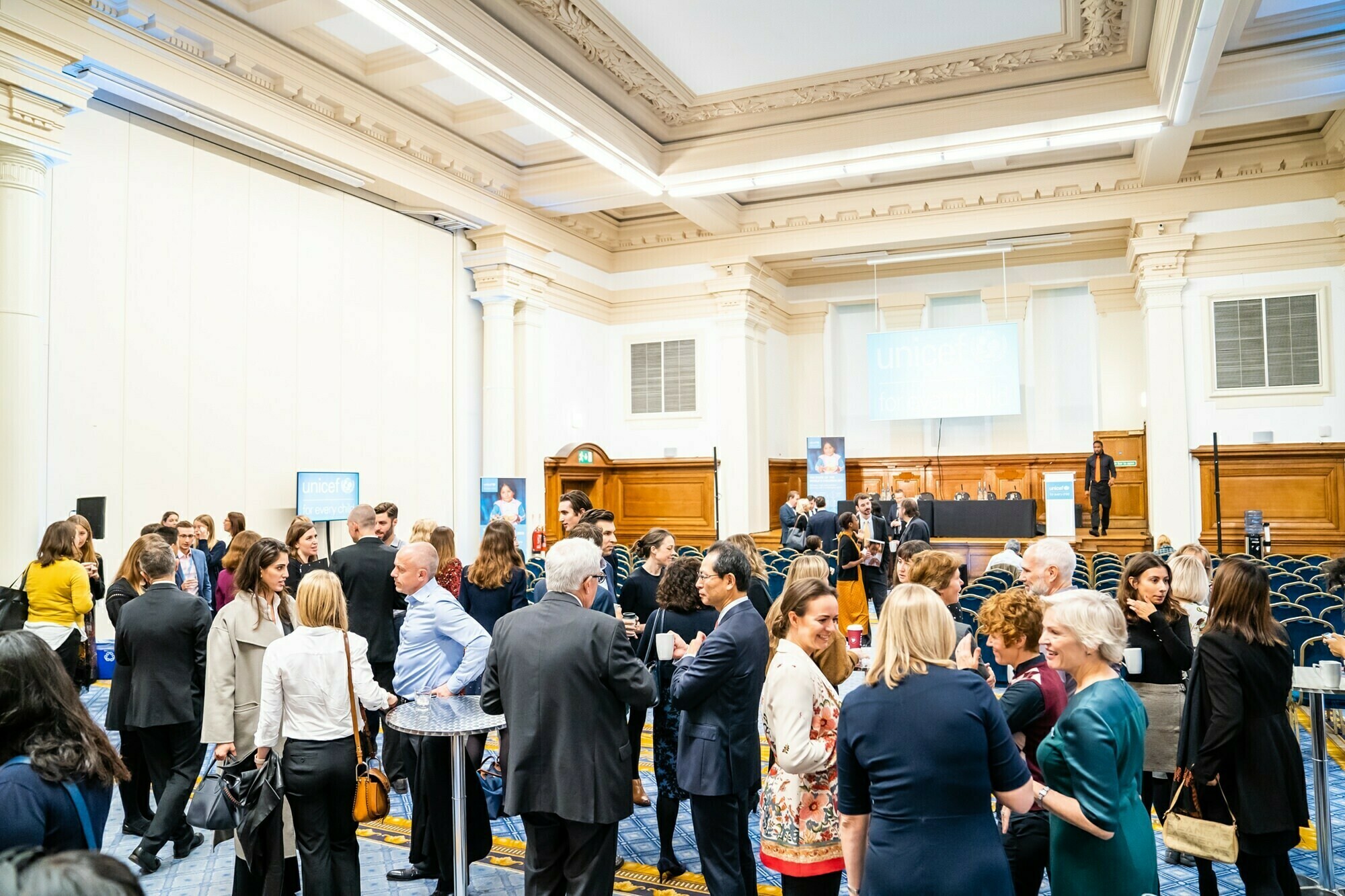 A large group of people in business attire gather and converse in a spacious, well-lit conference hall. Rows of empty chairs and a stage with a lectern are visible in the background, along with conference banners. James Gifford-Mead Photography - Event Photographer London