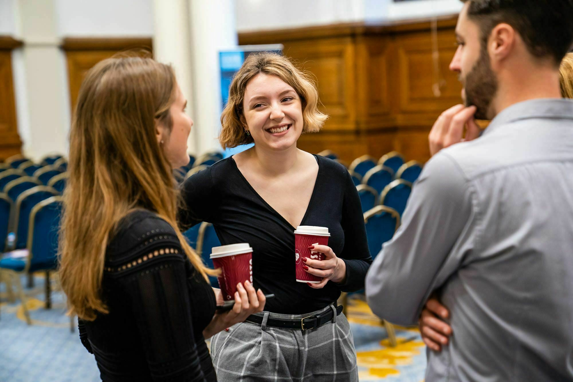 Three people are standing and talking in a room with rows of empty chairs. The woman in the center is smiling and holding a coffee cup. The other two people are also holding cups and engaged in conversation. They appear to be at an event. James Gifford-Mead Photography - Event Photographer London