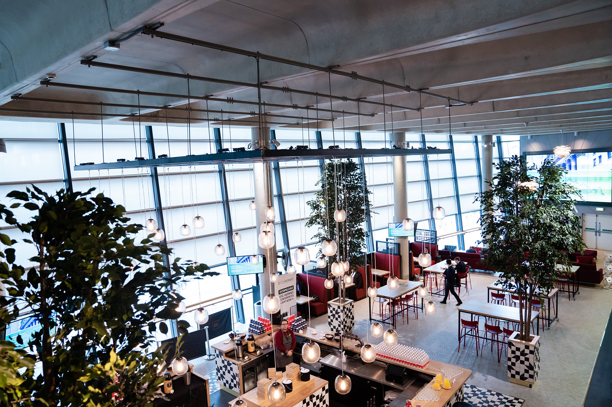 Interior view of a modern airport lounge with high ceilings and large windows. The space features hanging spherical lights, tall plants, red seating, and tables. Two people are walking near a counter area with black and white checkered details. James Gifford-Mead Photography - Event Photographer London