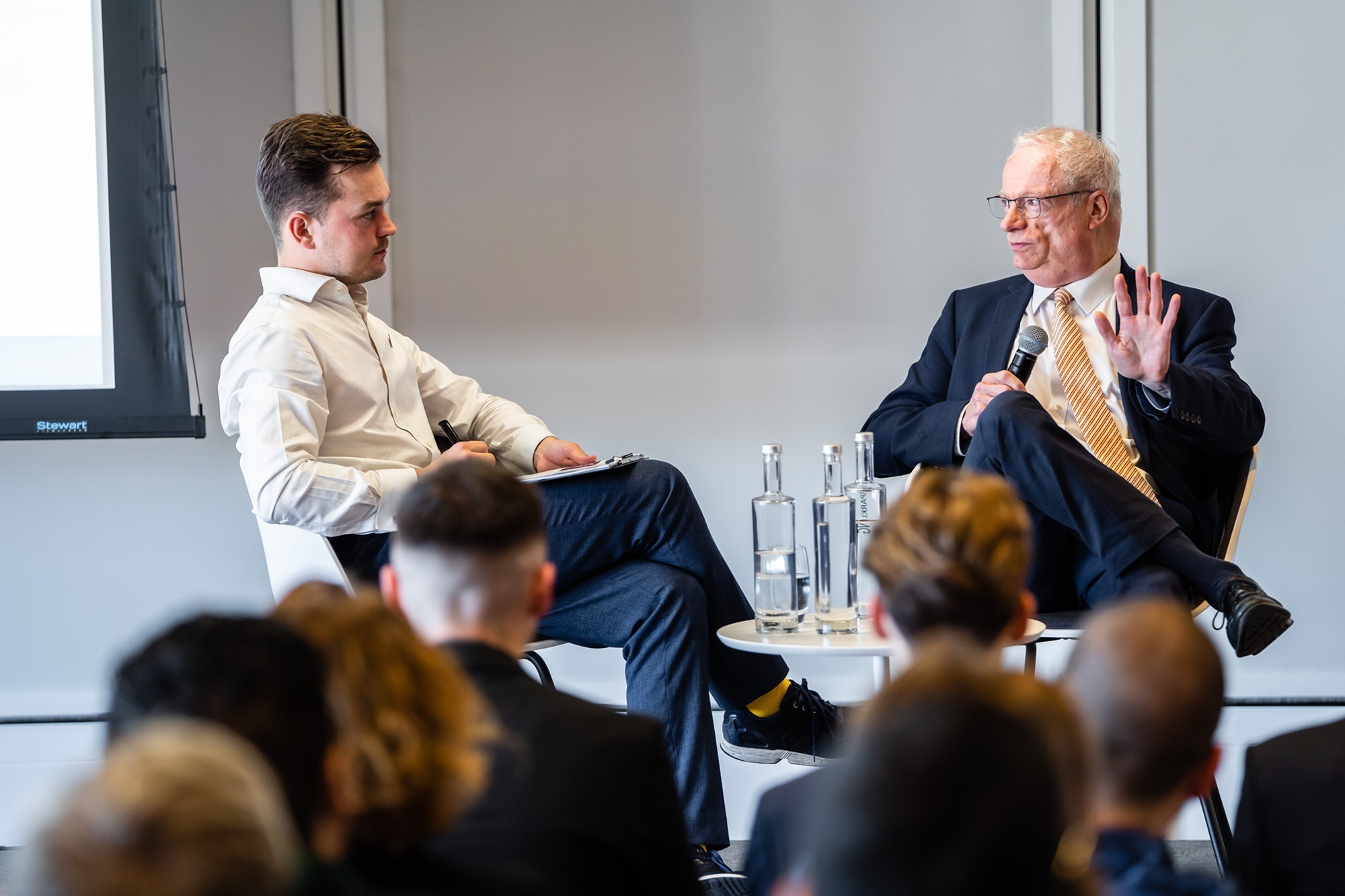 Two men are seated and engaged in conversation at a conference. The man on the left holds a notepad, wearing a white shirt and dark pants. The man on the right, in a suit and tie, gestures while speaking. Audience members are visible in the foreground. James Gifford-Mead Photography - Event Photographer London