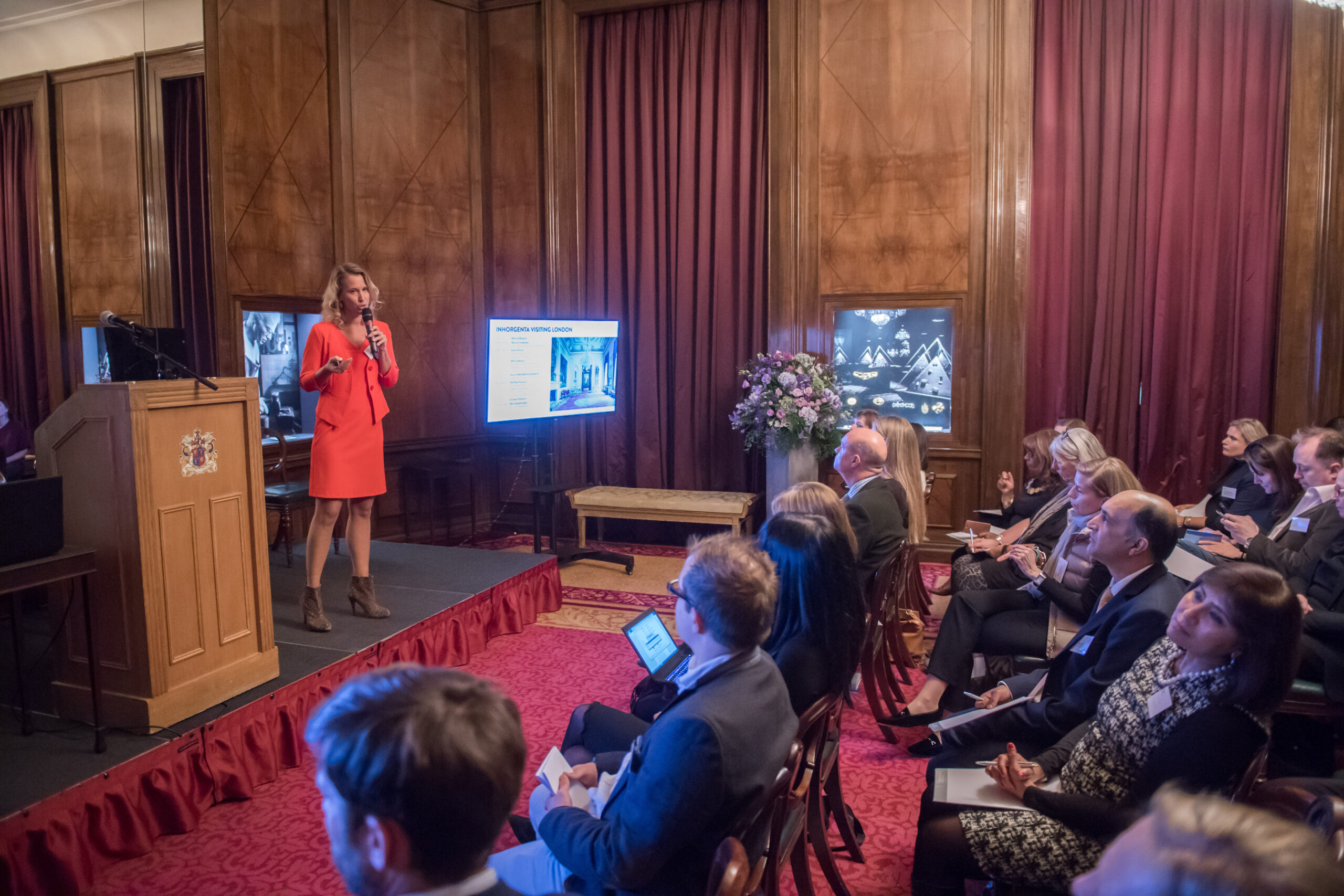 A woman in an orange dress is speaking at a podium in a richly decorated room with wooden paneling and red curtains. An audience is seated, listening attentively. A display screen shows a presentation slide next to the speaker. James Gifford-Mead Photography - Event Photographer London