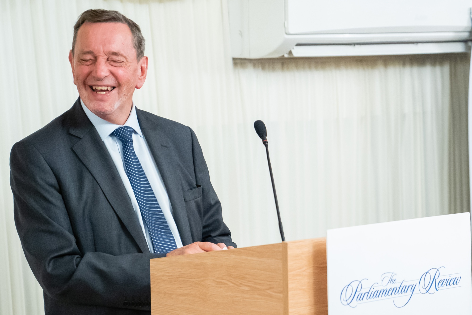 A man in a suit and tie is smiling while standing at a wooden podium with the sign "The Parliamentary Review." A microphone is attached to the podium. The background features light-colored drapes and an air conditioning unit. James Gifford-Mead Photography - Event Photographer London