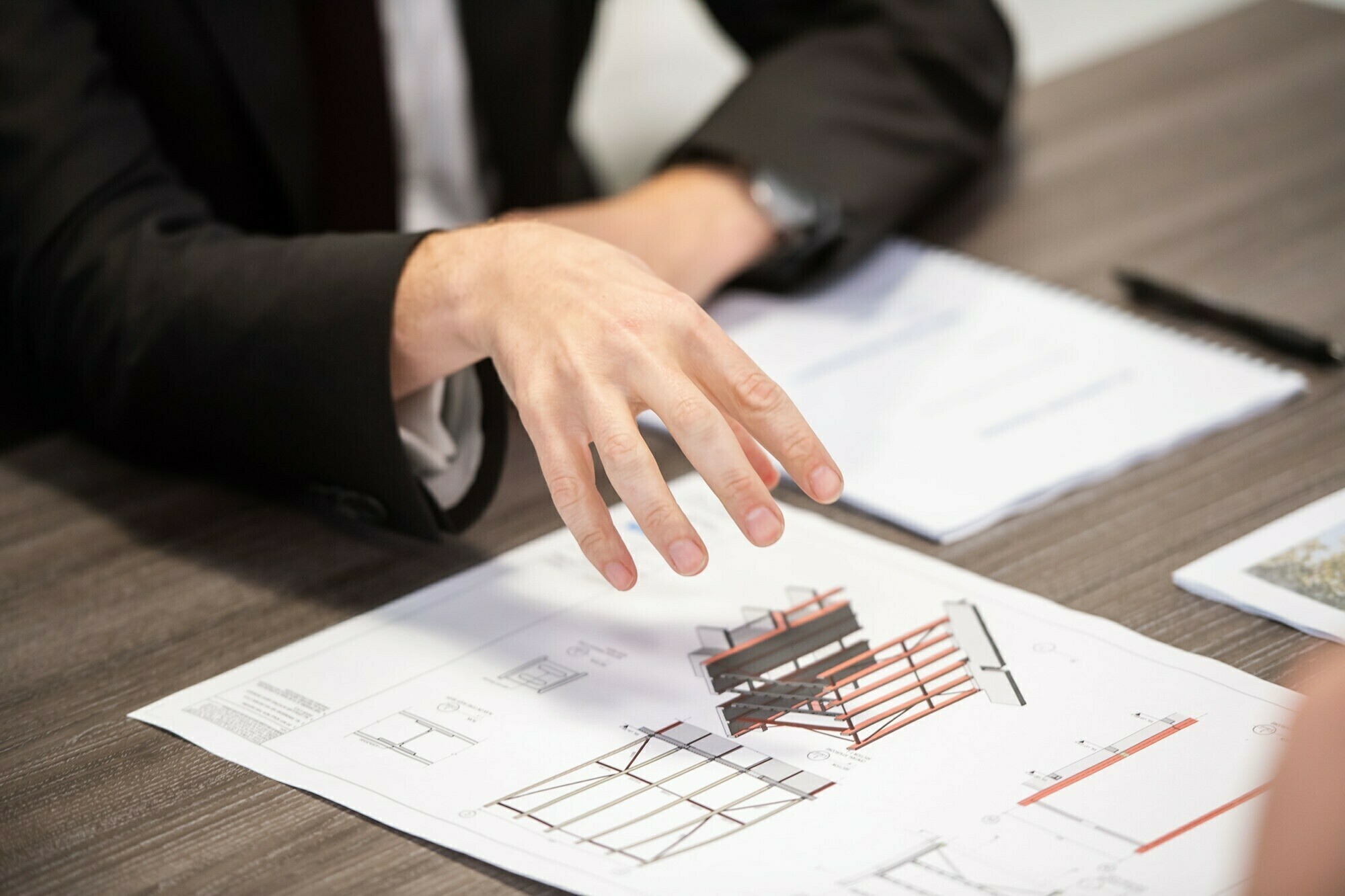 A person in a suit gestures over architectural plans spread on a wooden table. Various diagrams and designs are visible. A notebook and pen are placed nearby. James Gifford-Mead Photography - Event Photographer London