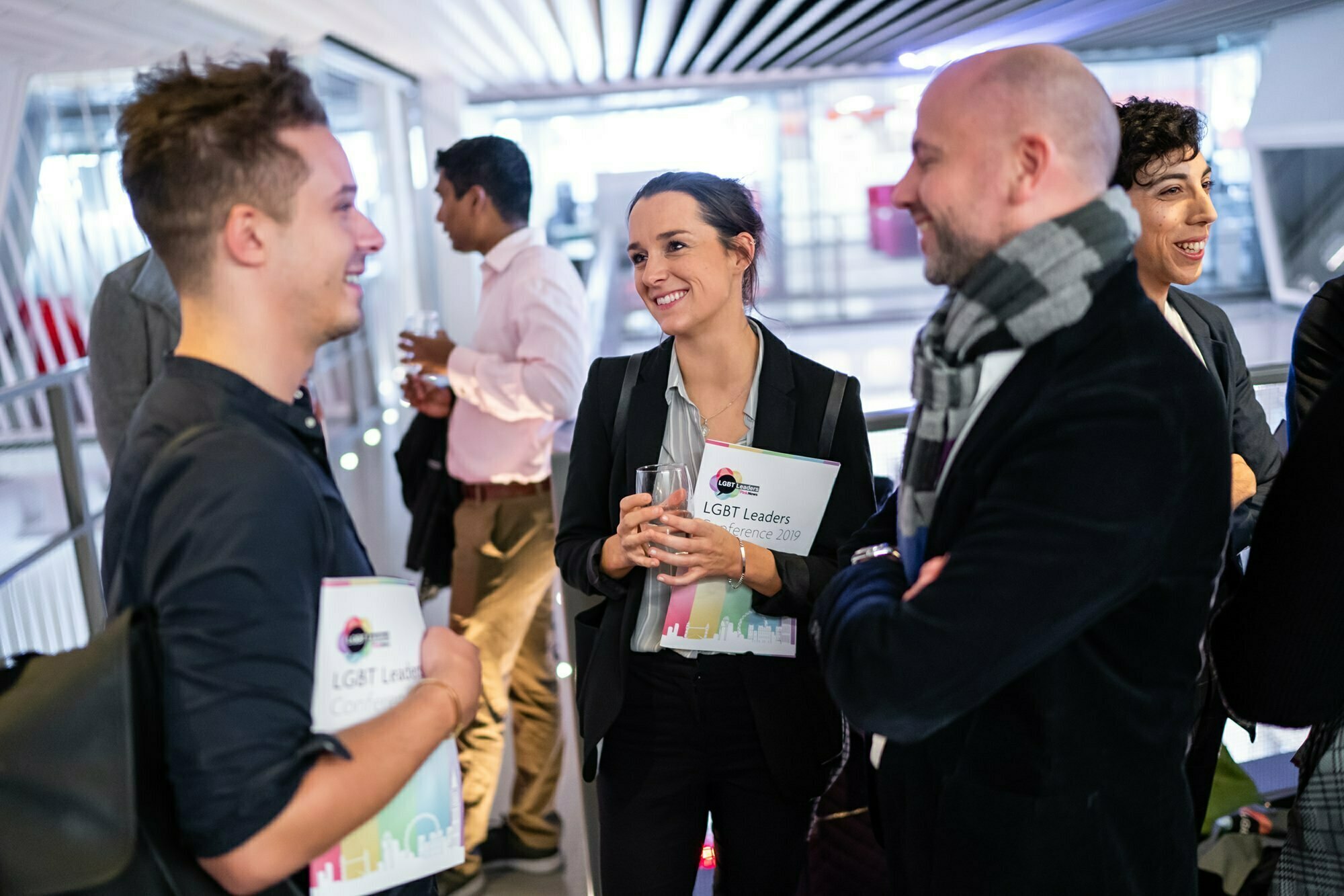 A group of professionally-dressed people are conversing at an event. They hold materials labeled "LGBT Leaders" and smile while engaged in discussion. The background features modern architecture with a bright, open ceiling. James Gifford-Mead Photography - Event Photographer London