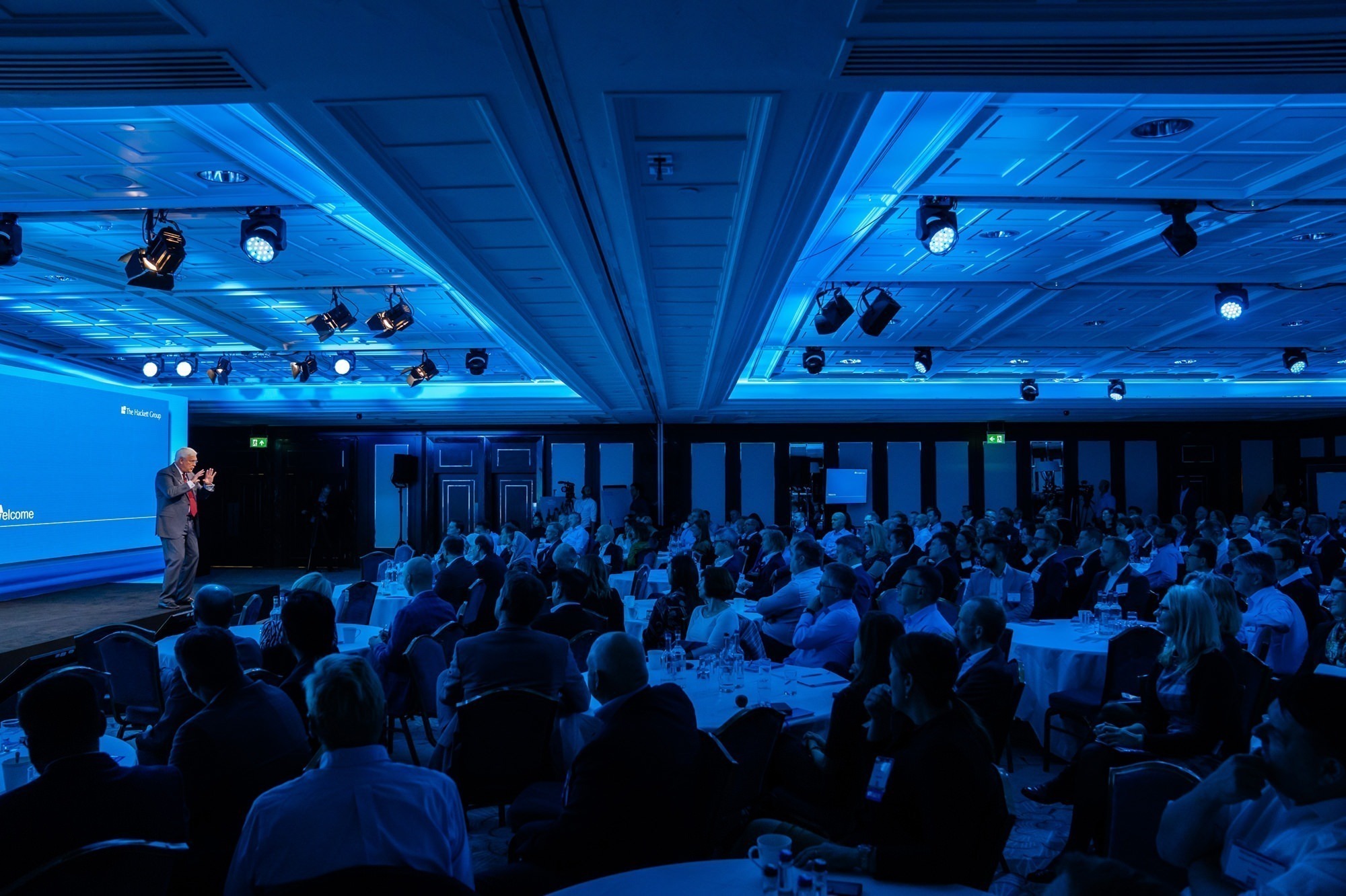 A speaker addresses a large audience in a dimly lit conference room. The room is bathed in blue light, with rows of seated attendees facing the stage. The ceiling has a grid pattern, and several round tables are arranged throughout the space. James Gifford-Mead Photography - Event Photographer London