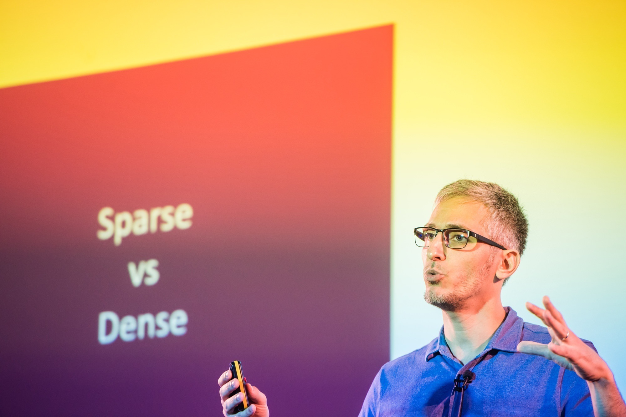 A person wearing glasses and a blue shirt speaks and gestures during a presentation. Behind them is a slide with the text "Sparse vs Dense" against a gradient background. James Gifford-Mead Photography - Event Photographer London