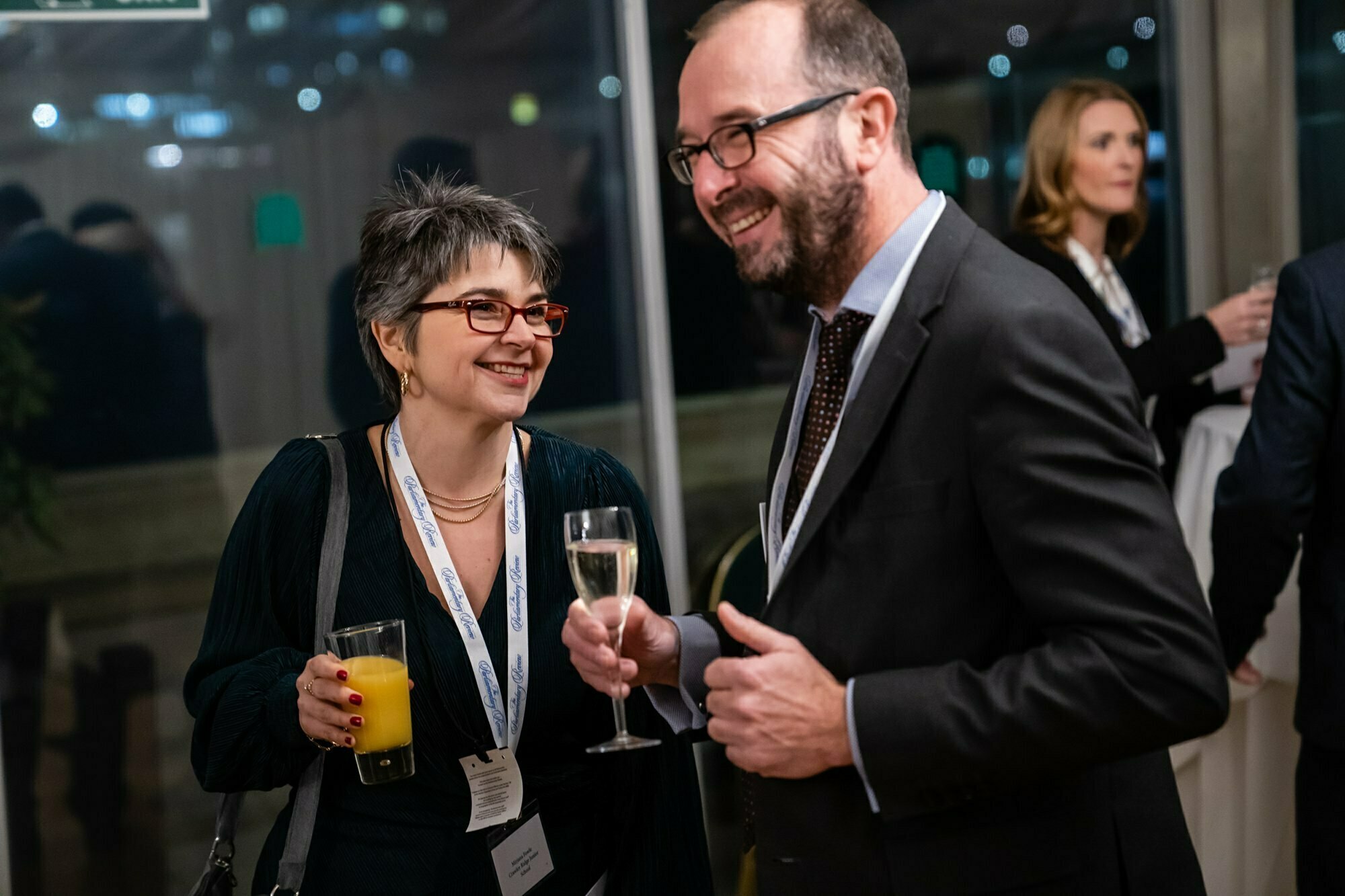 A woman and a man are engaging in conversation at an event. The woman is holding a glass of orange juice, and the man is holding a glass of champagne. Both are wearing lanyards and smiling. Other attendees are visible in the background. James Gifford-Mead Photography - Event Photographer London