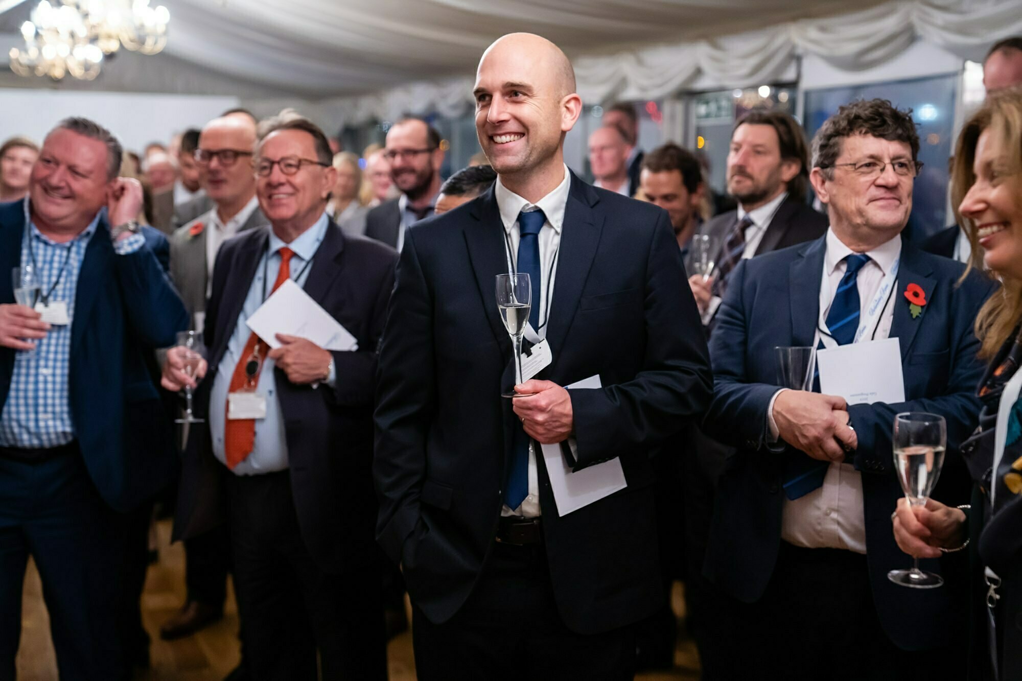A group of well-dressed people at a formal event, smiling and holding wine glasses. In the foreground, a bald man in a suit smiles broadly. The background features white draped decor and a chandelier. James Gifford-Mead Photography - Event Photographer London