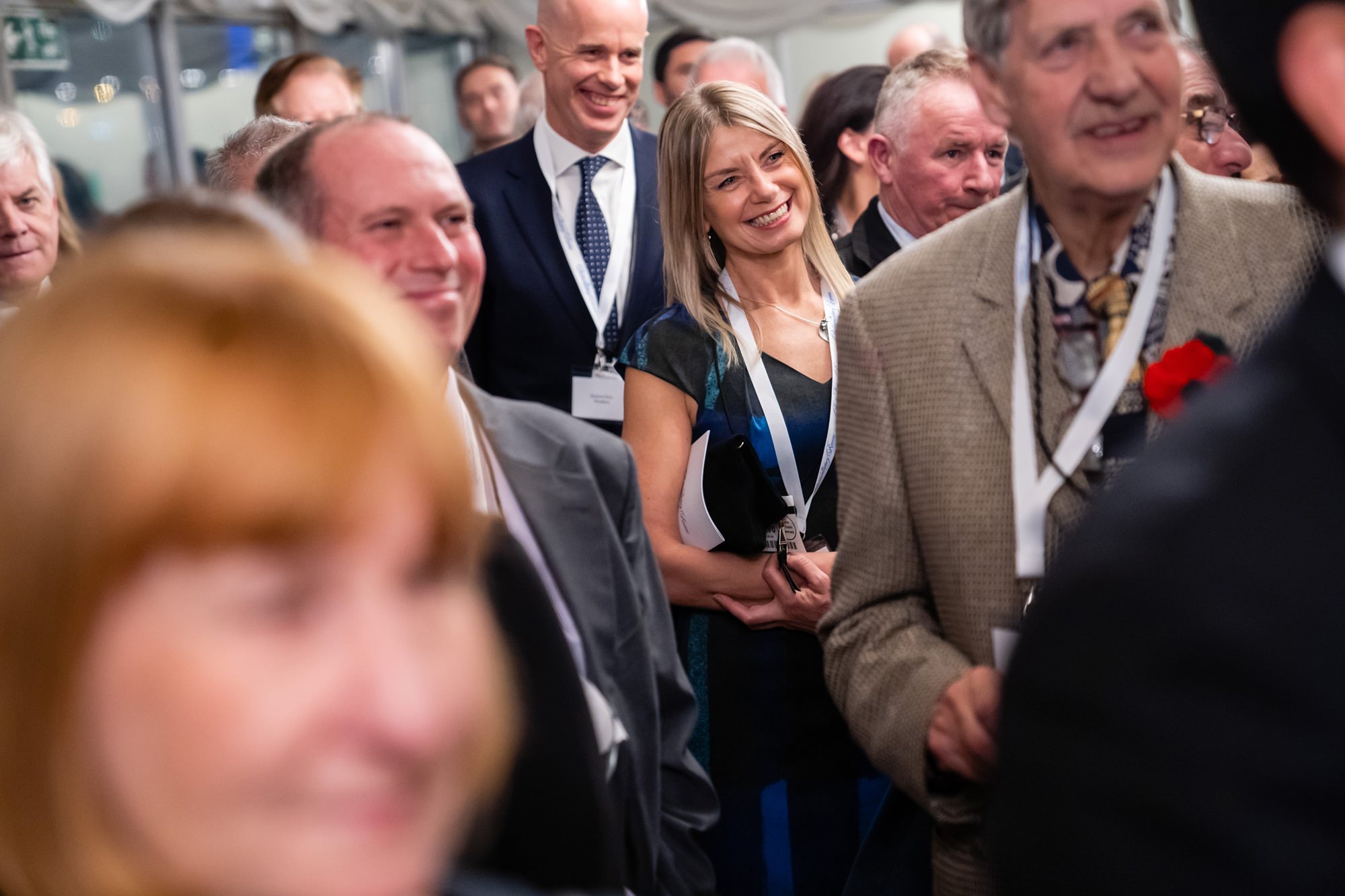 A group of professionally dressed people at a formal event, wearing name tags. They are smiling and seem engaged, with one woman in a blue dress and a man in a suit noticeable in the crowd. The setting is indoors. James Gifford-Mead Photography - Event Photographer London