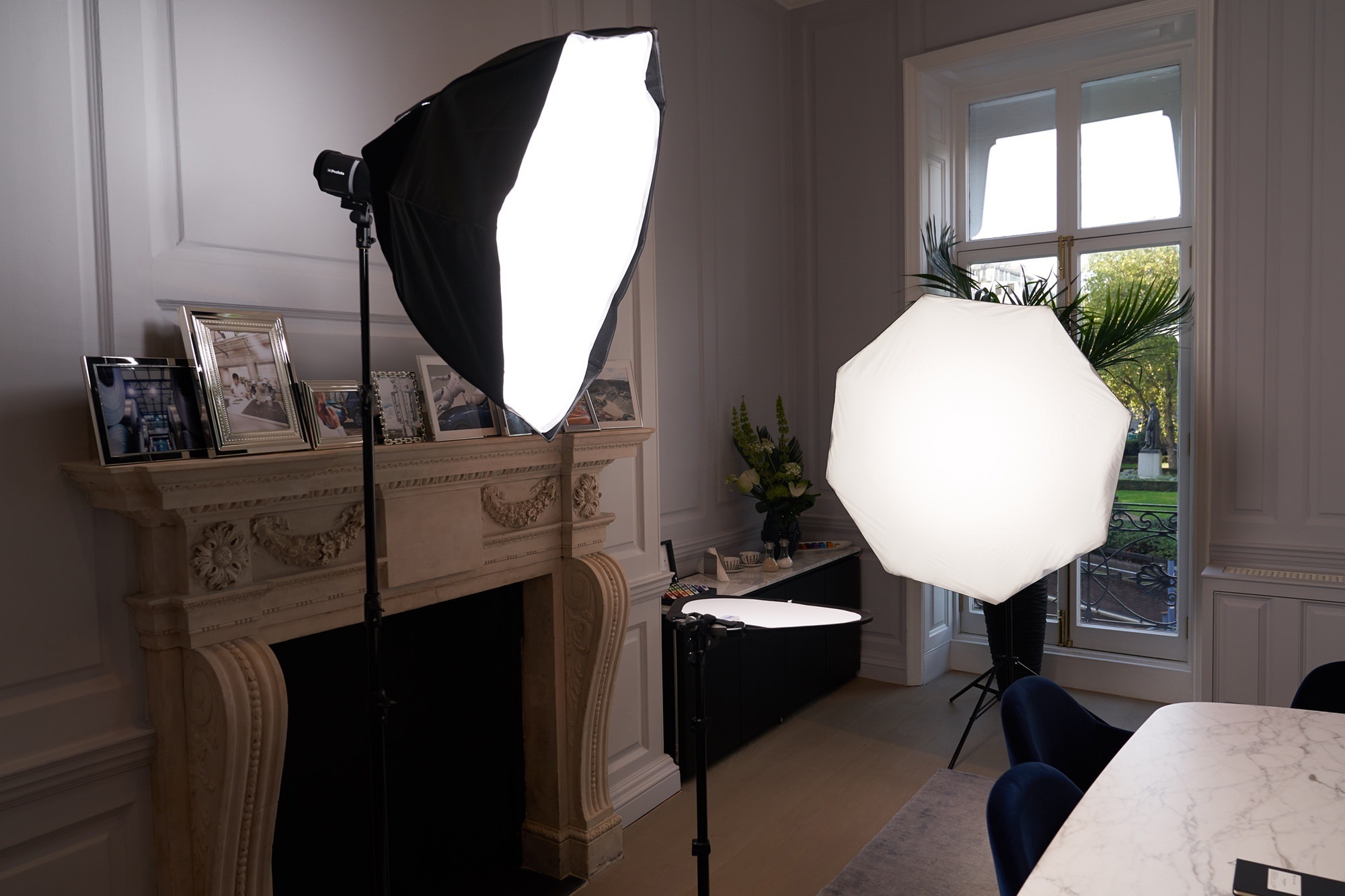 Photography lighting setup in a room with softboxes placed near a fireplace. Framed photos are on the mantel, and a large window offers a view of greenery outside. A table with chairs is partially visible in the foreground. James Gifford-Mead Photography - Event Photographer London