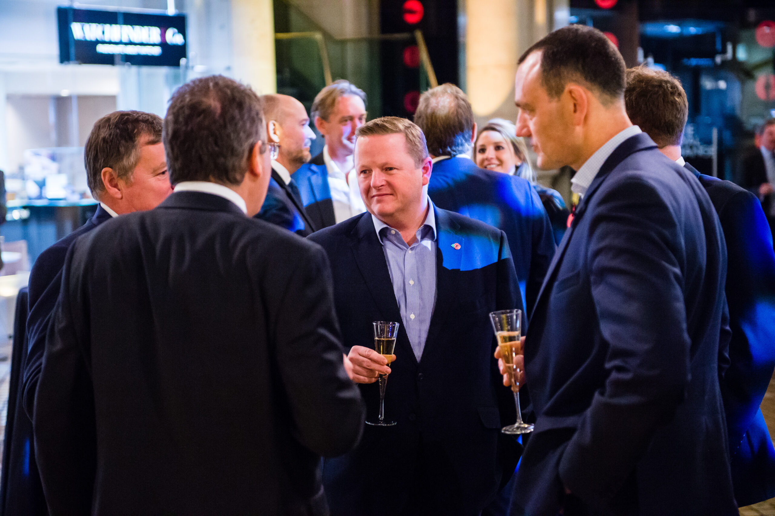 A group of people in business attire are gathered at a formal event, holding champagne flutes and engaging in conversation. The setting appears to be indoors, with modern decor and ambient lighting. James Gifford-Mead Photography - Event Photographer London