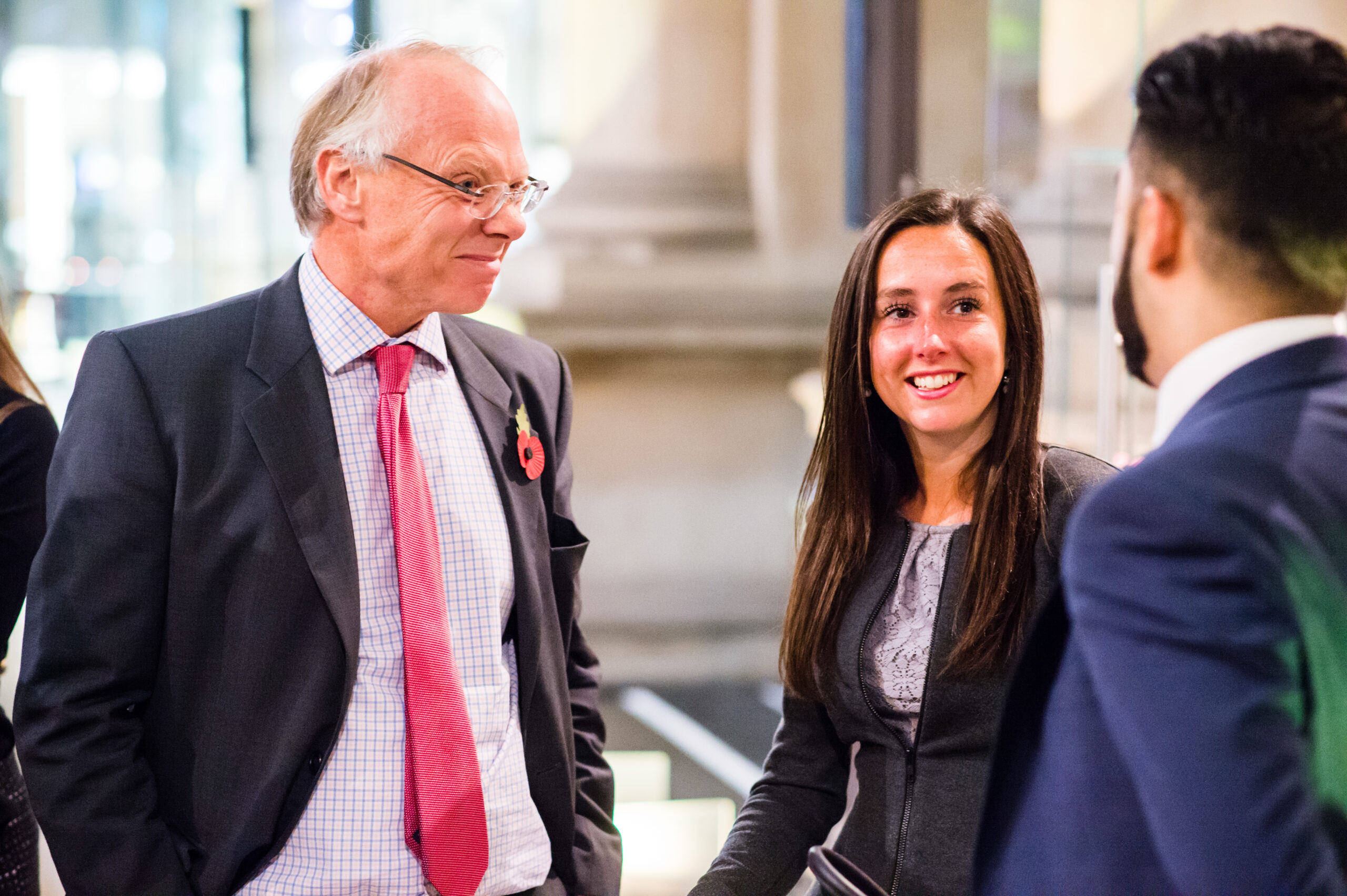 A group of four people, two men in suits with ties and two women in professional attire, are engaged in conversation indoors. One man has a poppy pin on his suit. They are standing in a well-lit area with blurred background elements. James Gifford-Mead Photography - Event Photographer London