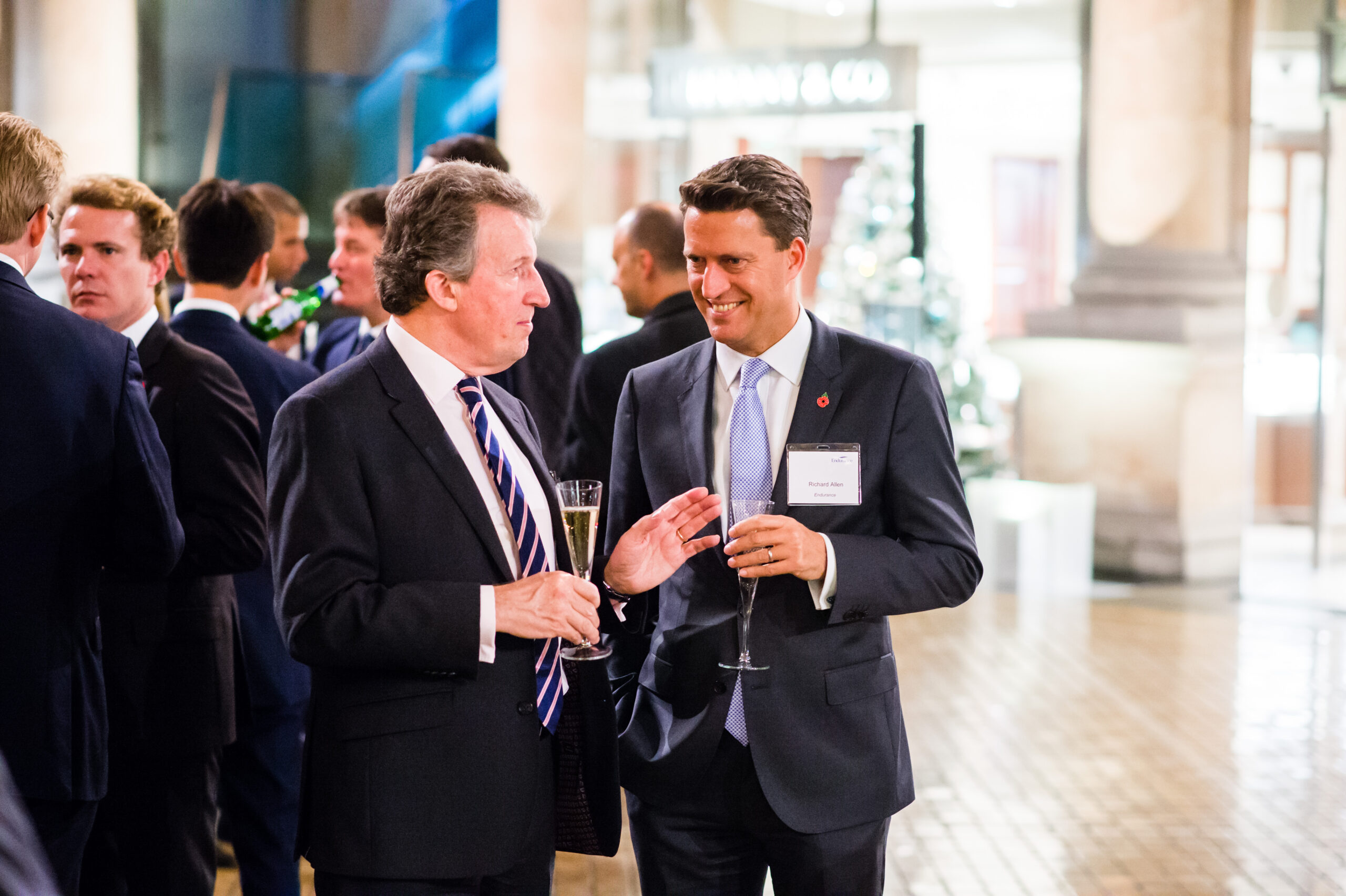 Two men in suits are conversing and holding glasses at a formal event. They are standing amidst a group of people in a well-lit venue, with one man gesturing as he speaks. A sign and a Christmas tree are visible in the background. James Gifford-Mead Photography - Event Photographer London