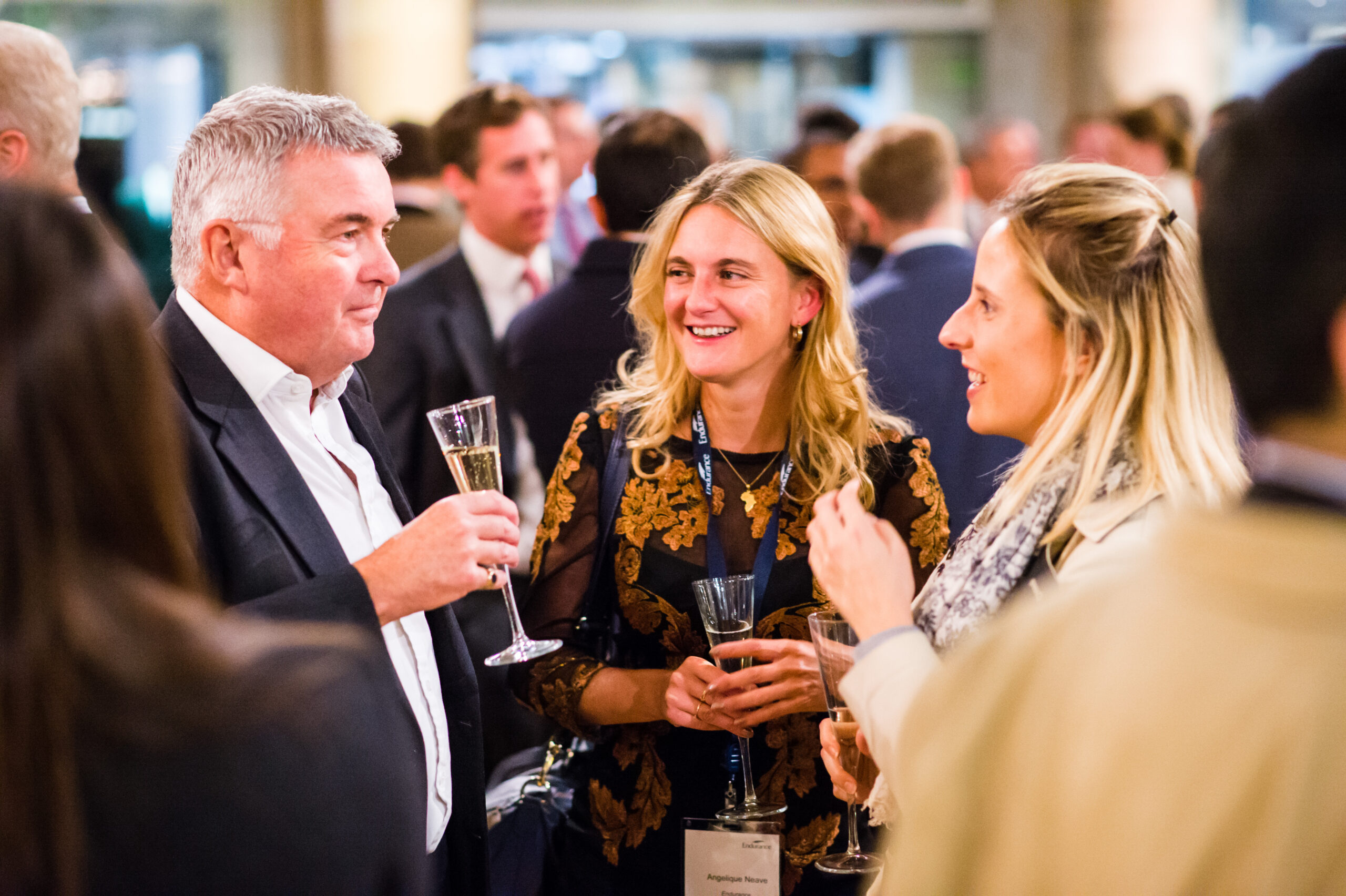 A group of people in formal attire are socializing at an indoor event. They are holding glasses of champagne and smiling, with a blurred background of more people engaged in conversation. James Gifford-Mead Photography - Event Photographer London