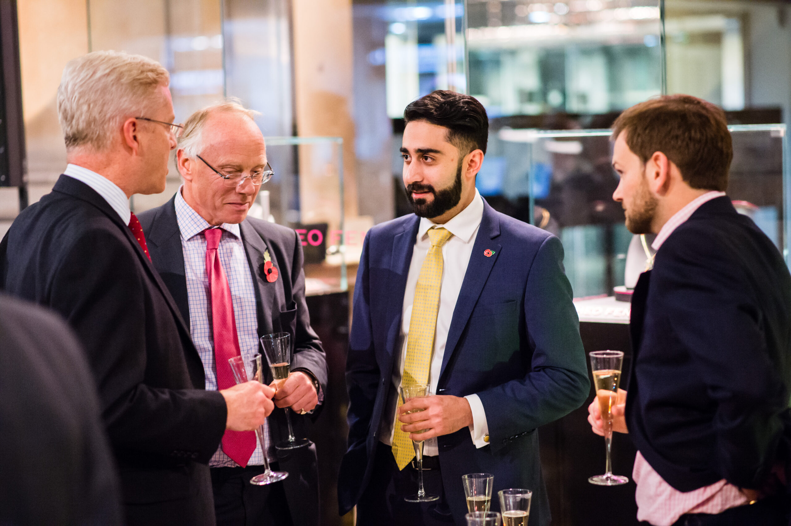 Four men in business attire stand in a group, holding champagne glasses. They are engaged in conversation at what appears to be a formal event or reception. Display cases are visible in the background. James Gifford-Mead Photography - Event Photographer London