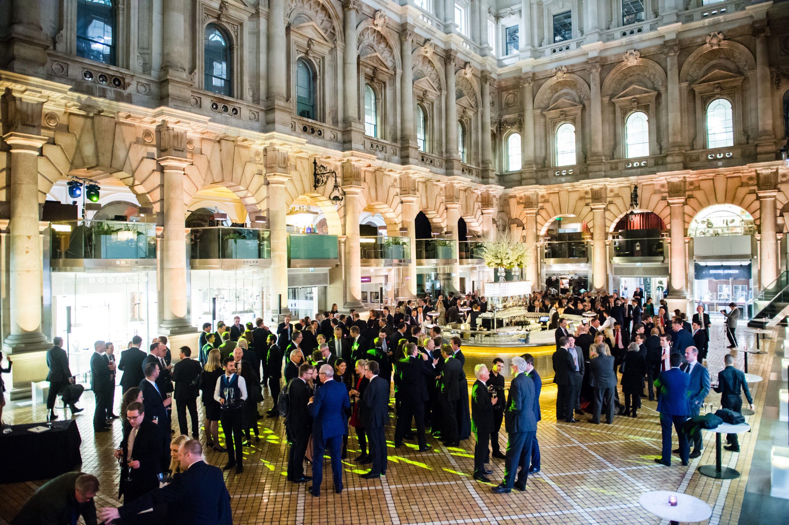 A large group of people in formal attire socializing at an elegant event inside a grand building with ornate arches, a high ceiling, and warm lighting. The floor is tiled, and there is a central circular fountain. James Gifford-Mead Photography - Event Photographer London