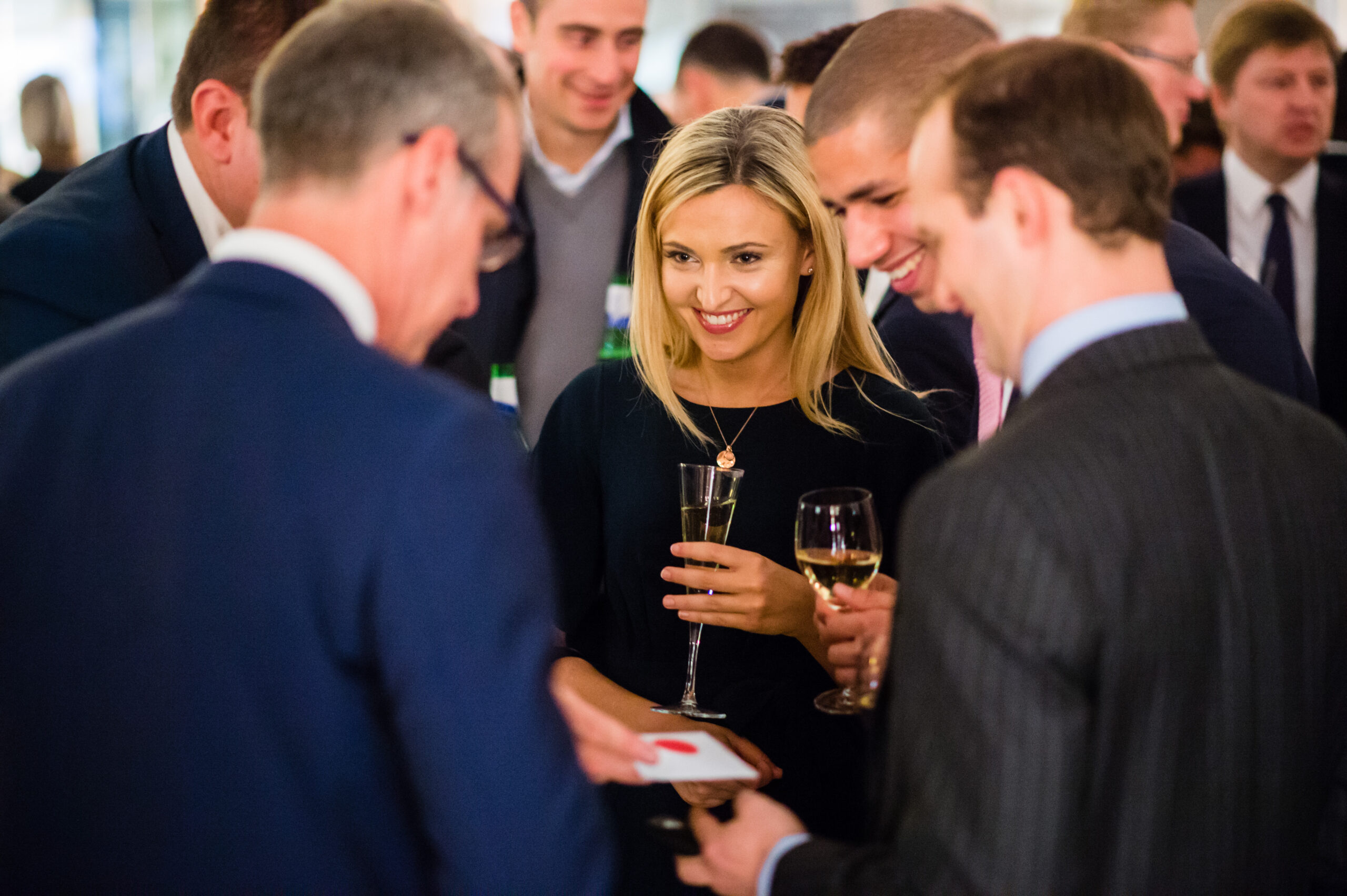 A group of people in formal attire are standing together, holding drinks and smiling. A man in the foreground holds playing cards, appearing to perform a magic trick. The setting suggests a social or networking event. James Gifford-Mead Photography - Event Photographer London