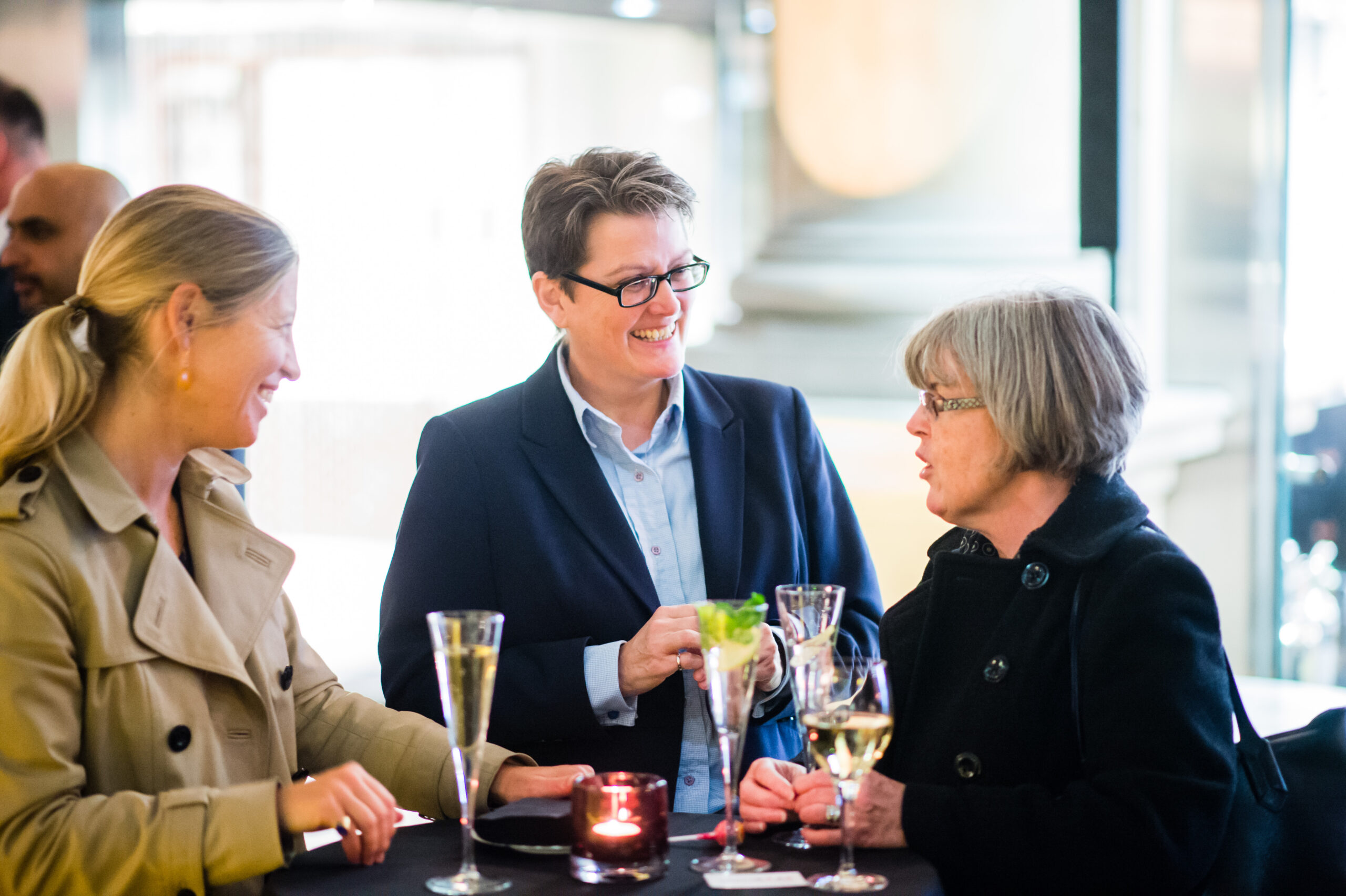 Three people stand around a table, smiling and talking. They are holding drinks, including champagne and a cocktail. The setting appears to be indoors, in a well-lit, formal environment. A candle is on the table. James Gifford-Mead Photography - Event Photographer London